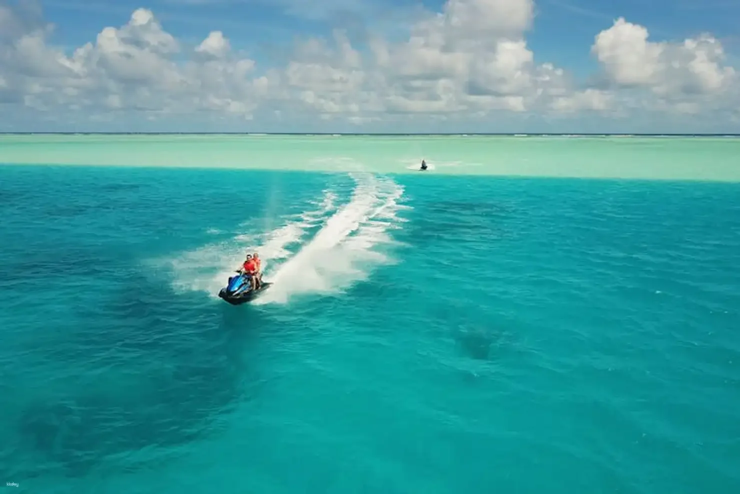 Person riding a jet ski on turquoise water, with a sandy barrier and a boat in the distance under a partly cloudy sky.
