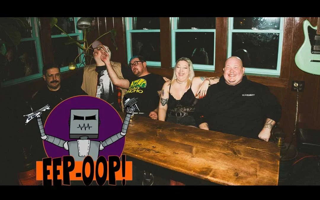 Five people sitting at a wooden table in a bar, with one person adjusting their glasses, smiling, and a green guitar hanging on the wall. There is a cartoon robot with the words "FEP OOP!" in the bottom left corner.
