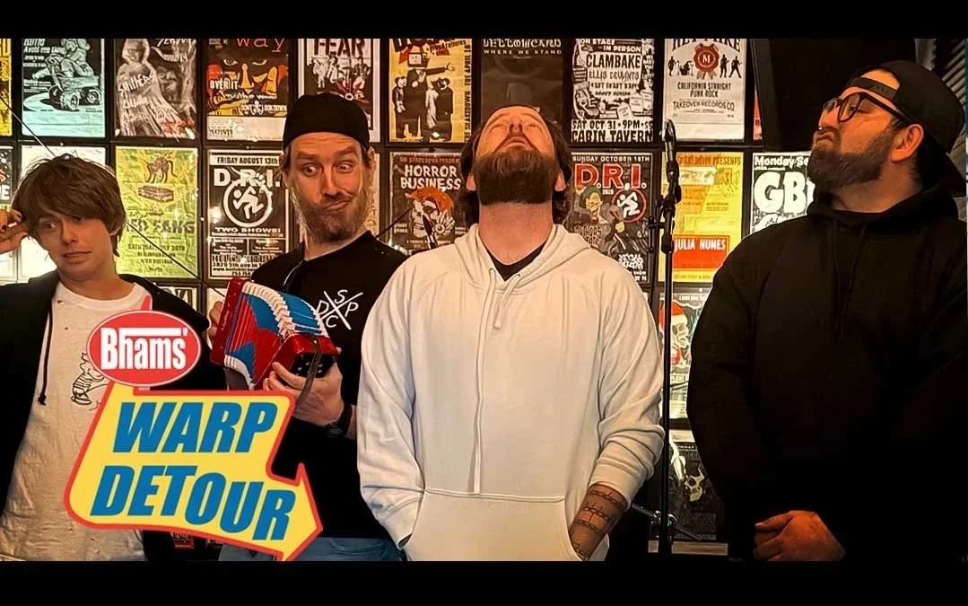 A group of four men standing in front of a wall covered with posters, some with creative and colorful designs. One man is holding a small Moog synthesizer, and another is wearing a black cap. They appear to be at an event or performance, with a promotional logo in the bottom left corner reading 'Bram's WARP DETOUR'.