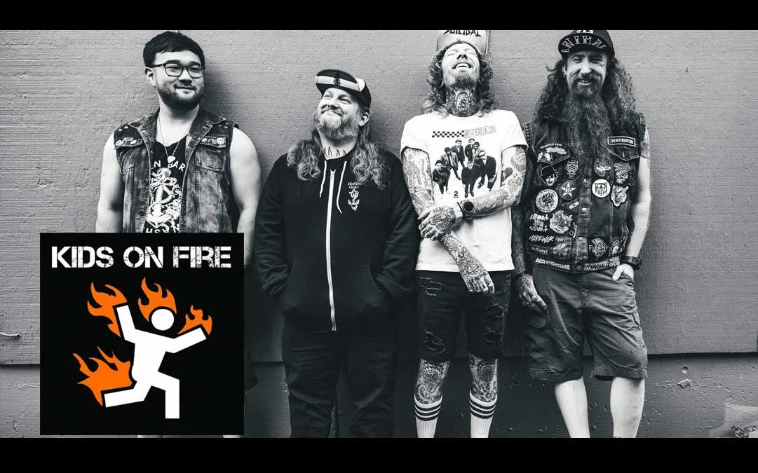 Four men standing against a wall with a Kids on Fire logo in the lower left corner. The men are dressed casually, with two wearing denim vests with patches, one with a hoodie, and one with tattoos, a graphic t-shirt, and shorts. They are smiling and relaxed.