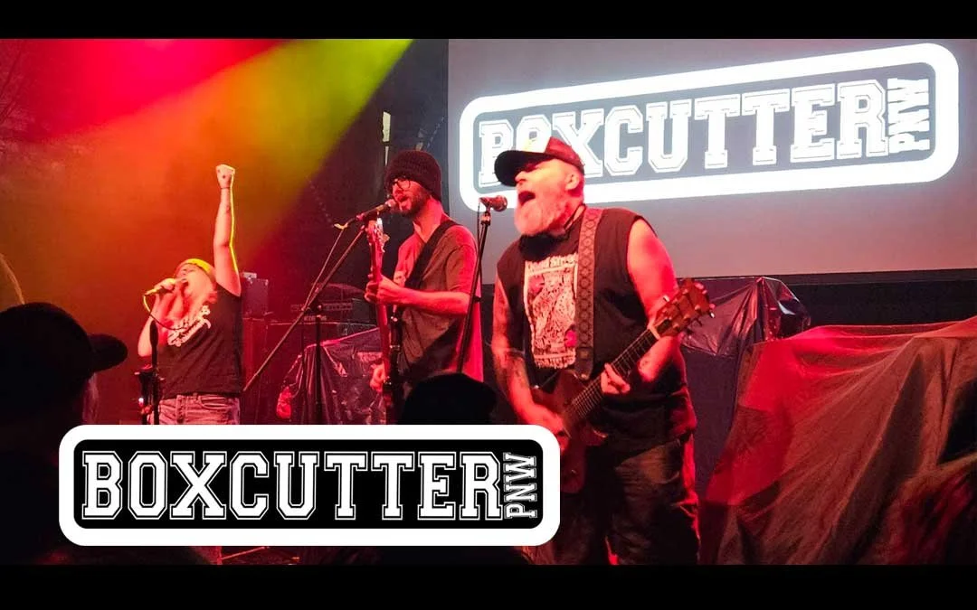 A band performing on stage with illuminated 'BOXCUTTER' sign in the background; a female vocalist singing with arm raised, a guitarist wearing glasses and beanie, and a bearded man in a sleeveless shirt playing guitar.