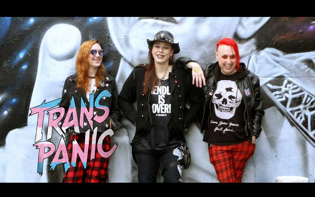 Three women with colorful hair and rocker fashion standing in front of a graffiti mural, smiling and laughing.