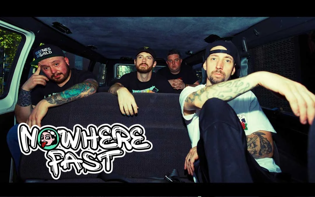A group of four men with tattoos sitting inside a vehicle, with a band logo "Nowhere Fast" on the seat in front of them.