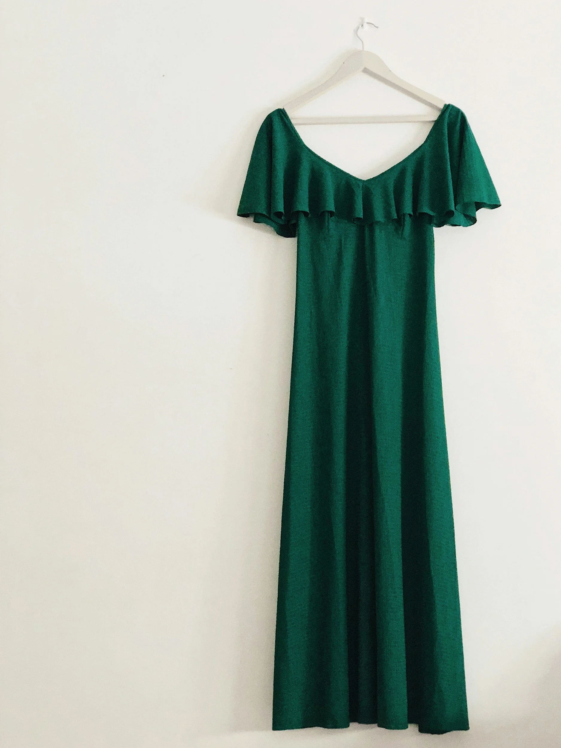 A long green dress with a ruffled off-the-shoulder neckline hanging on a white hanger against a plain white wall.