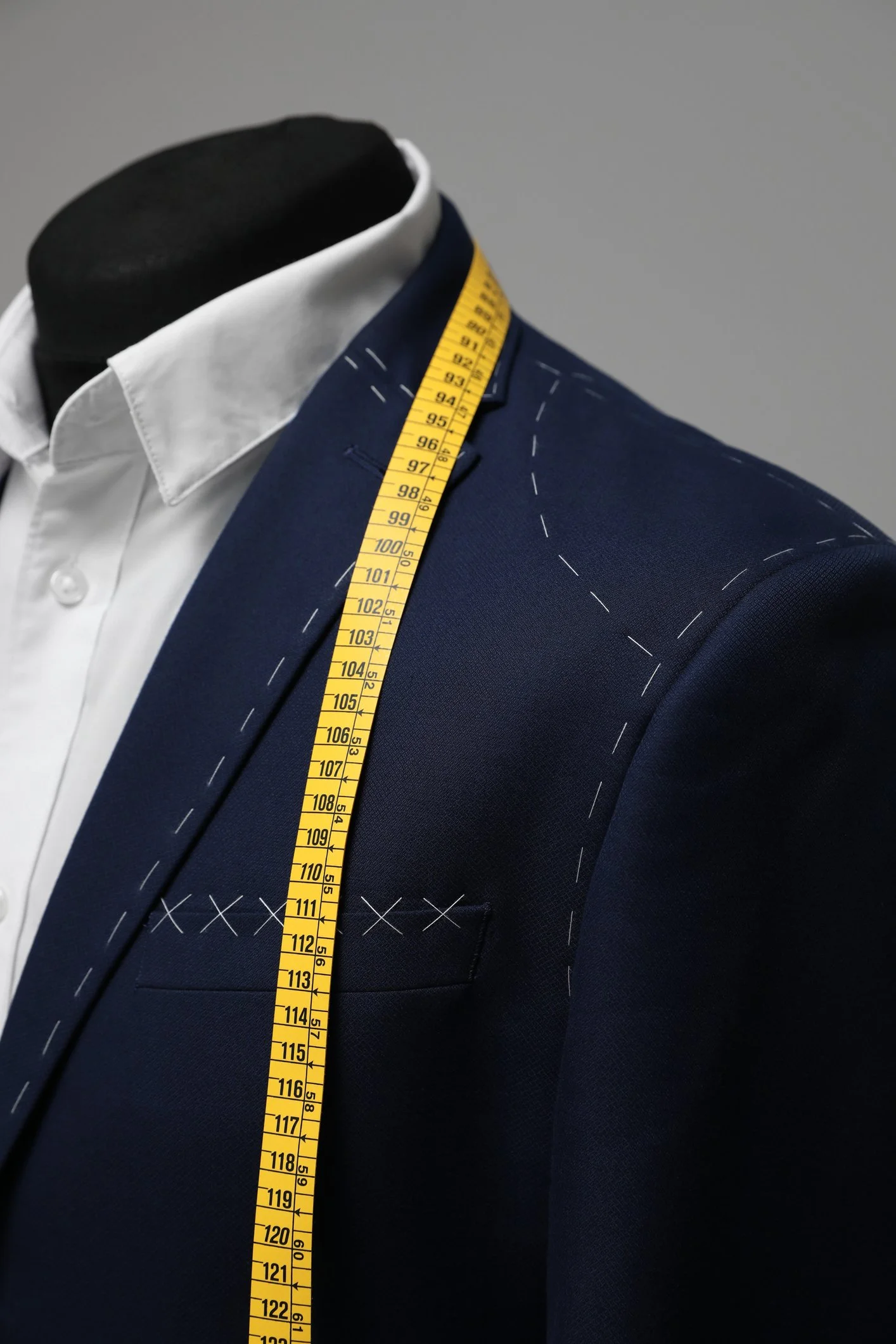 Close-up of a tailored navy blue suit jacket on a mannequin, with white stitching marks and a yellow measuring tape draped over the shoulder, next to a white dress shirt.