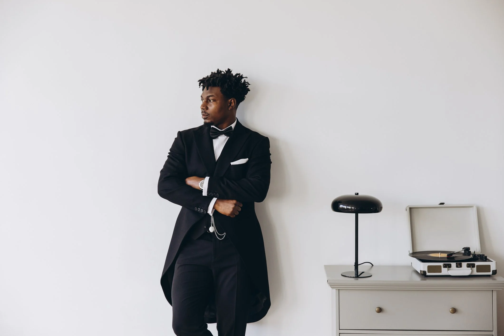 A man in a black tuxedo with a bow tie leaning against a plain white wall, next to a gray chest of drawers with a black vintage record player and a black modern table lamp.