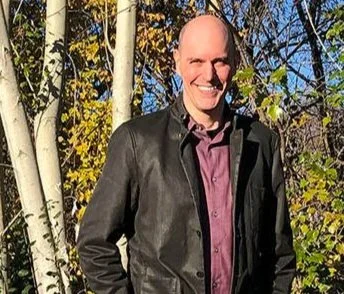 A smiling man standing outdoors among trees with yellow leaves.