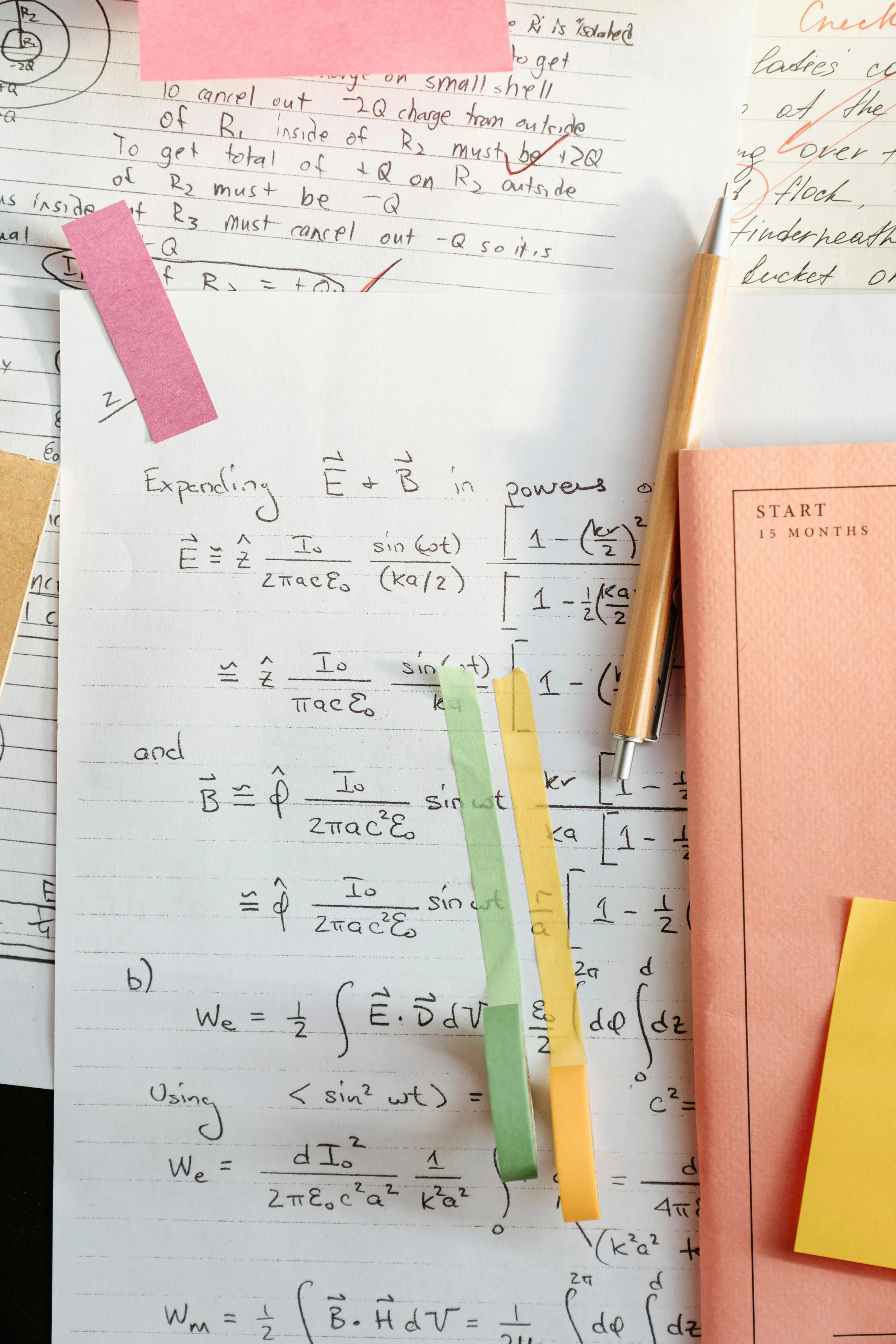 Close-up of a cluttered desk with handwritten notes, colored sticky notes, a pen, and a pink notebook on top of mathematical and scientific papers.