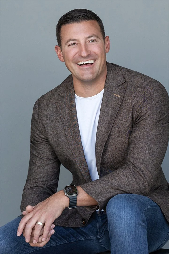 Professional portrait of Chris Kruseiwicz seated in a brown blazer and jeans against a gray studio background, photographed by Alex Abercrombie.
