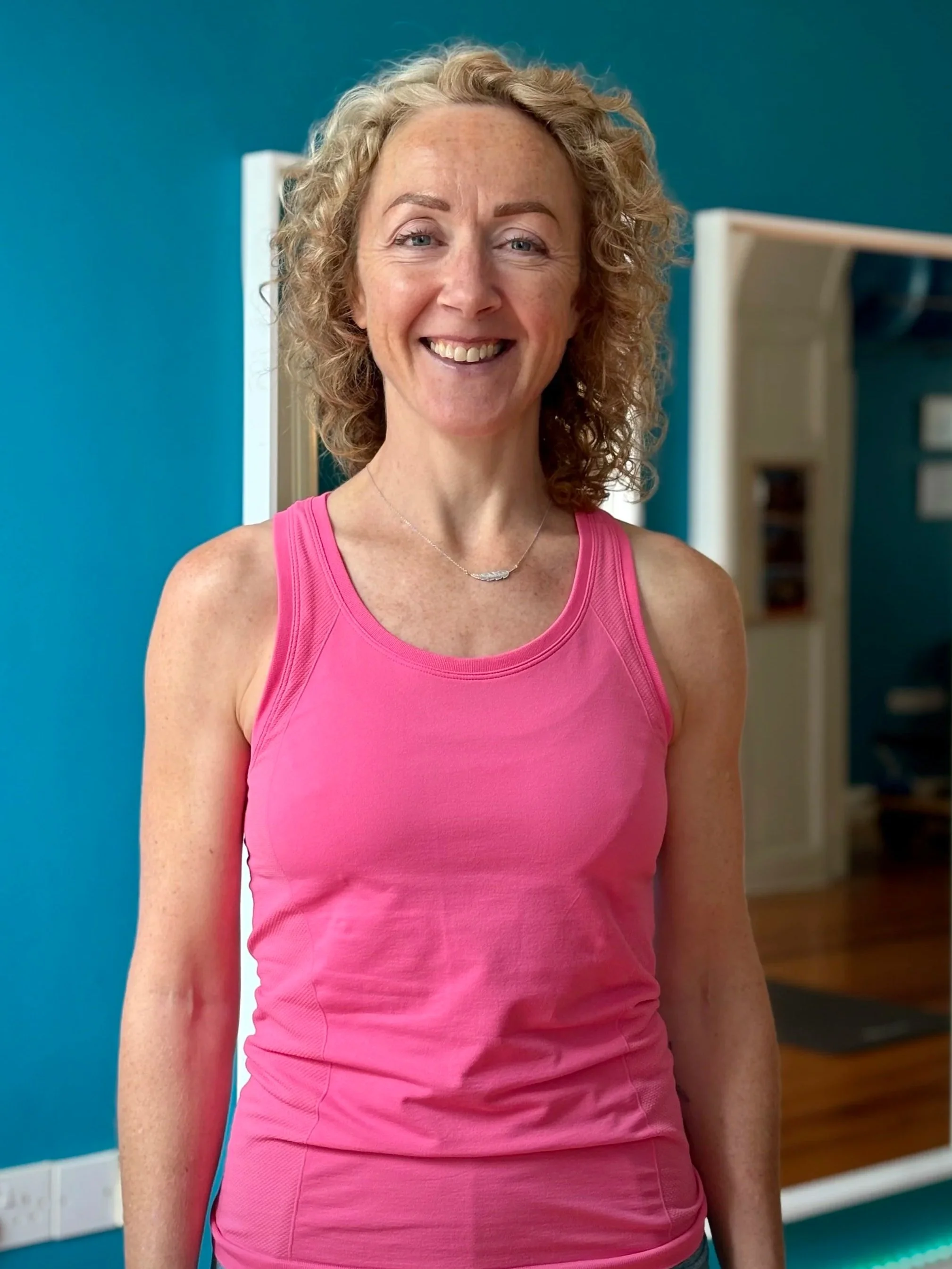 A smiling woman with dark hair holding a pink yoga mat in an indoor setting, wearing a white athletic tank top.