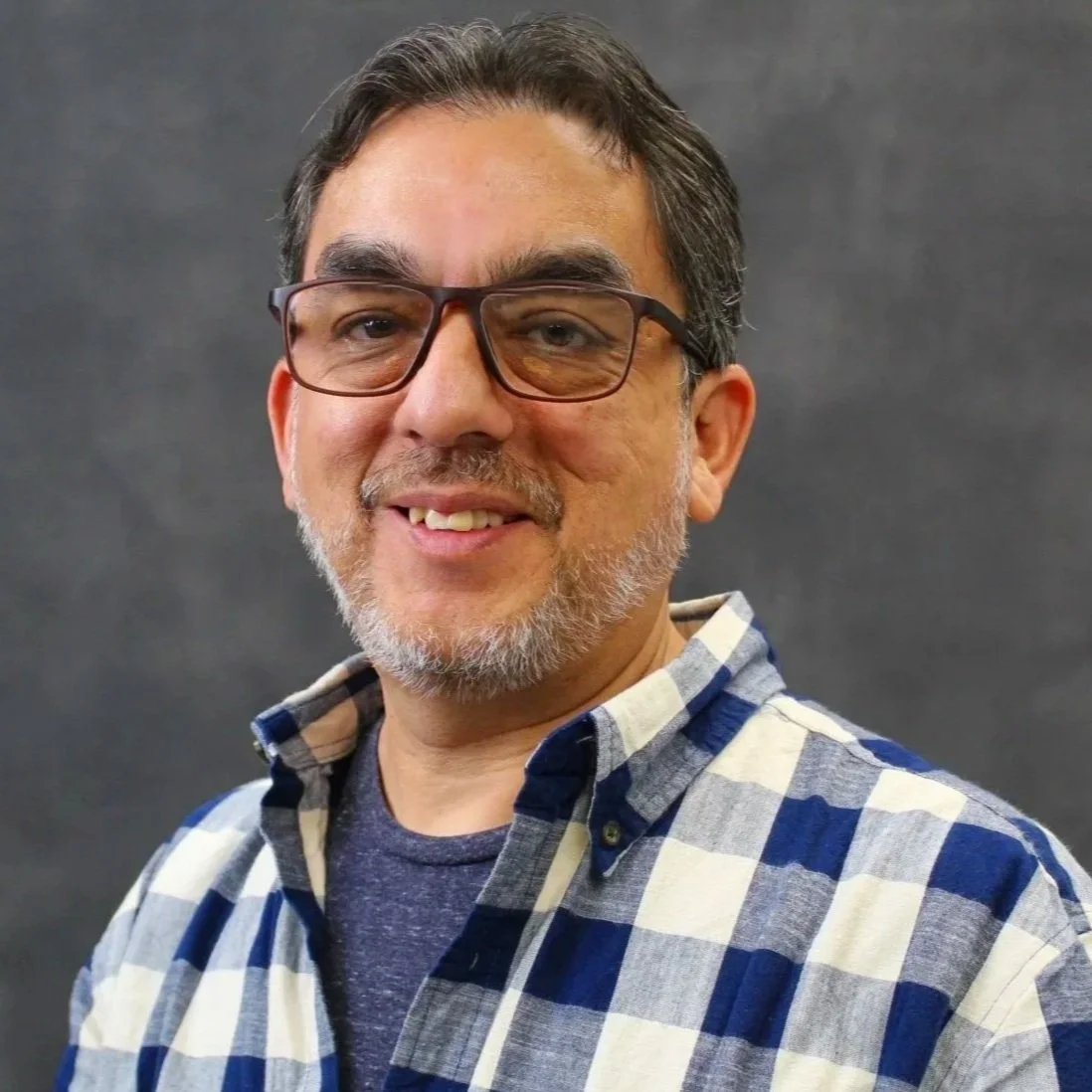 Portrait of a man with glasses, a beard, and gray hair, wearing a blue and white checkered shirt and smiling, against a dark background.