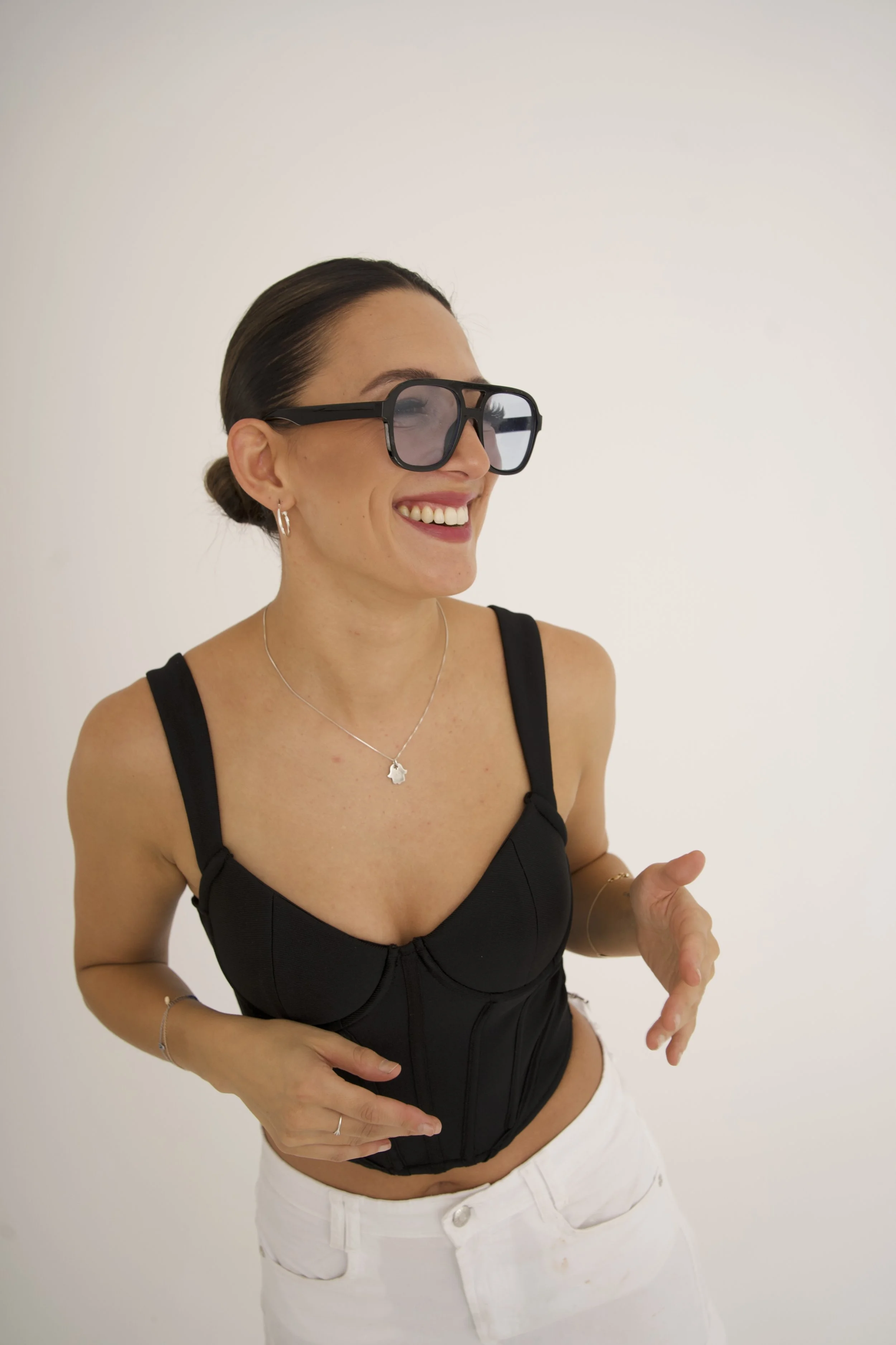 Ning-Ning is smiling, wearing large black sunglasses, black crop top, white pants, and silver jewelry, standing against a plain white background, for a DJ portrait.