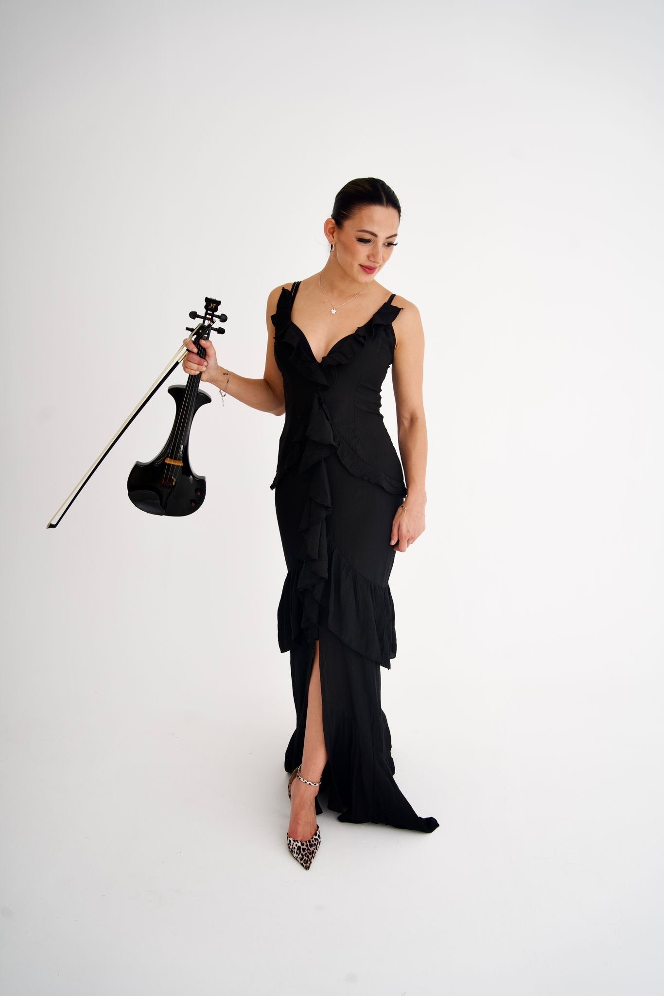 Ning-Ning wears a black ruffled dress holding a black electric violin and bow, standing against a plain white background, wearing leopard print high heels.