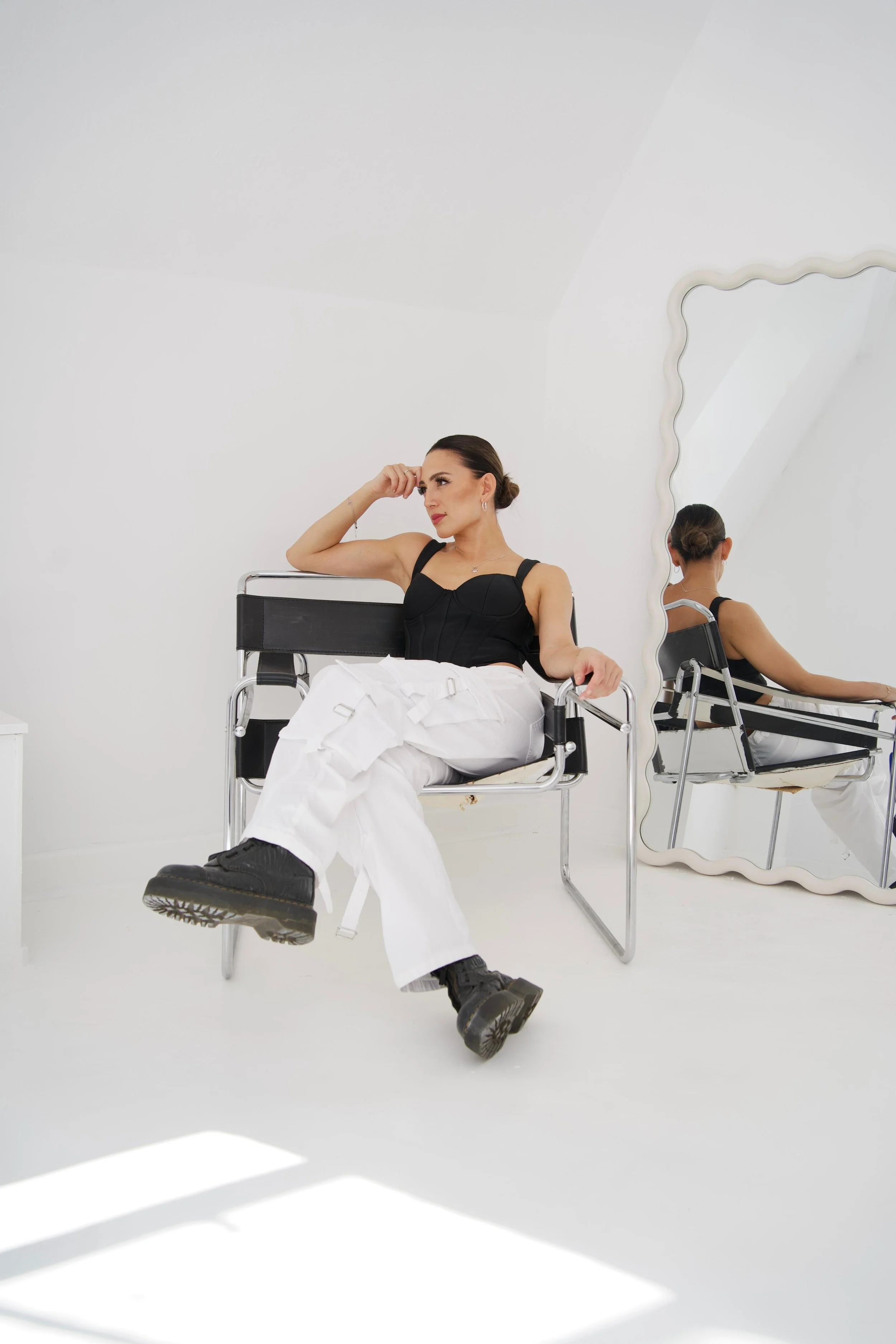 Ning-Ning sits on a black chair in a white room, looking thoughtfully to her left, with a large mirror reflecting her back. For a DJ portrait.