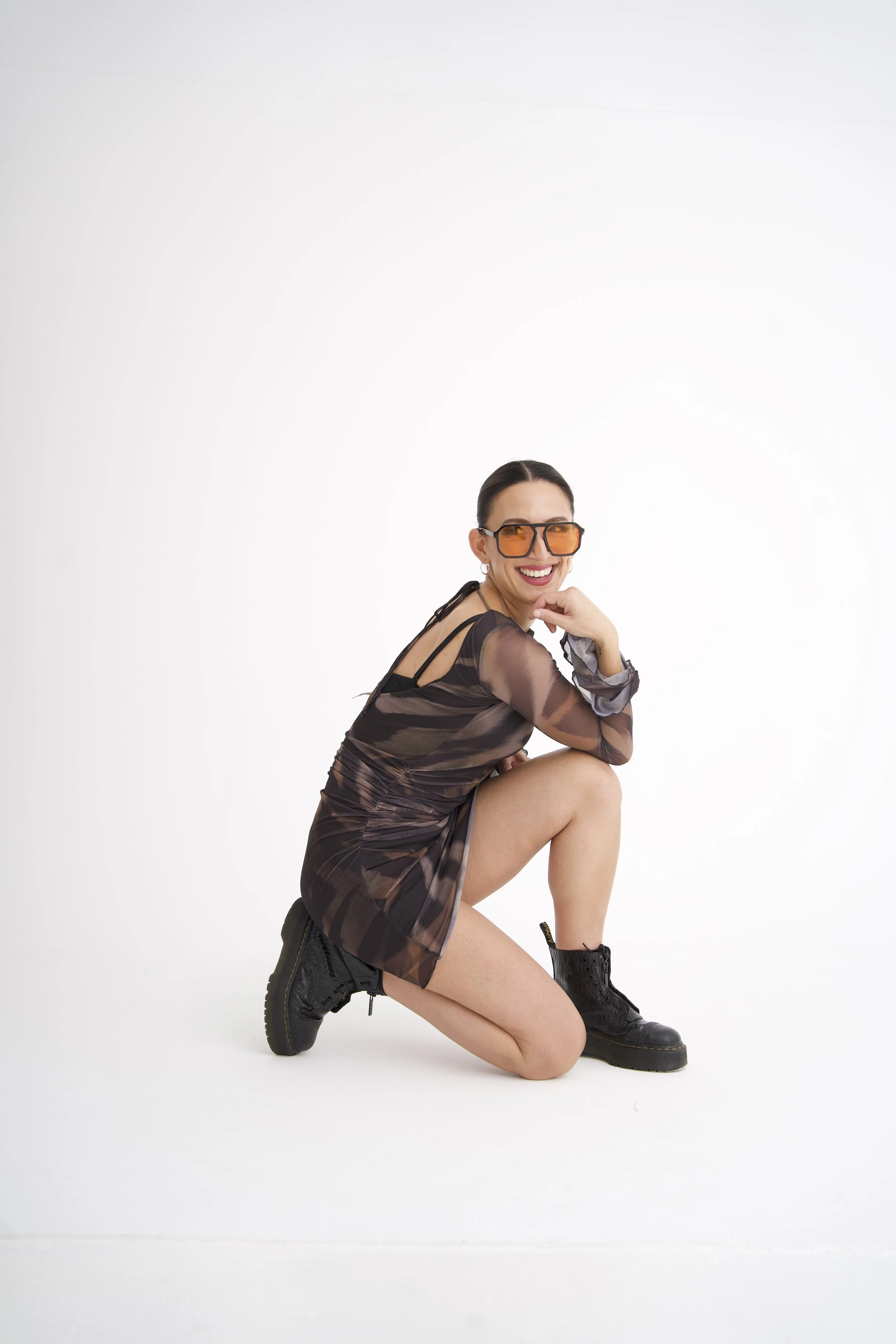 Ning-ning is wearing large orange sunglasses, a sheer brown dress, black combat boots, and black underwear, kneeling on one knee and smiling at the camera against a plain white background, for another DJ portrait.