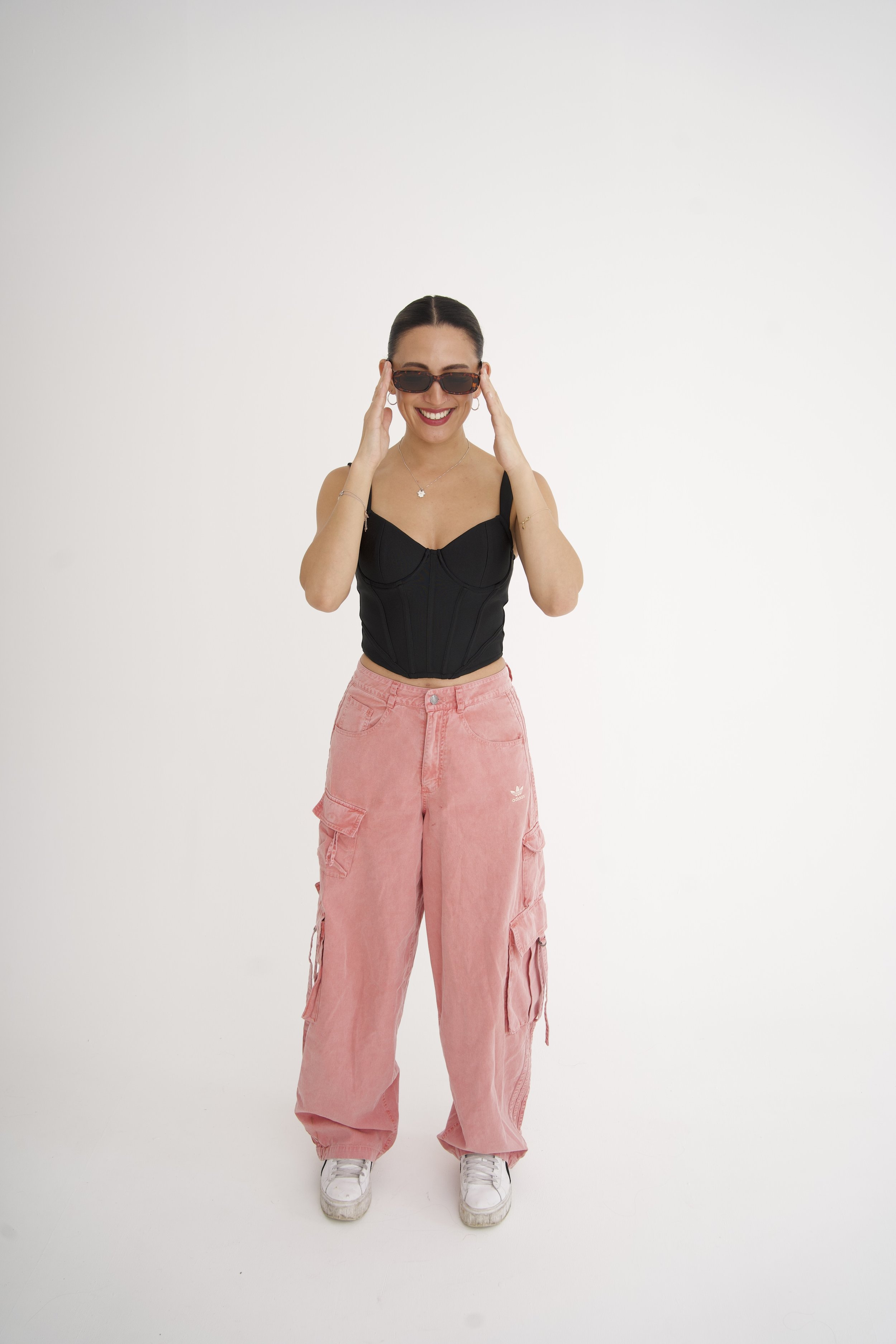 Ning-Ning is wearing sunglasses, a black top, pink cargo pants, and sneakers, smiling in front of a plain white background, for a DJ portrait.
