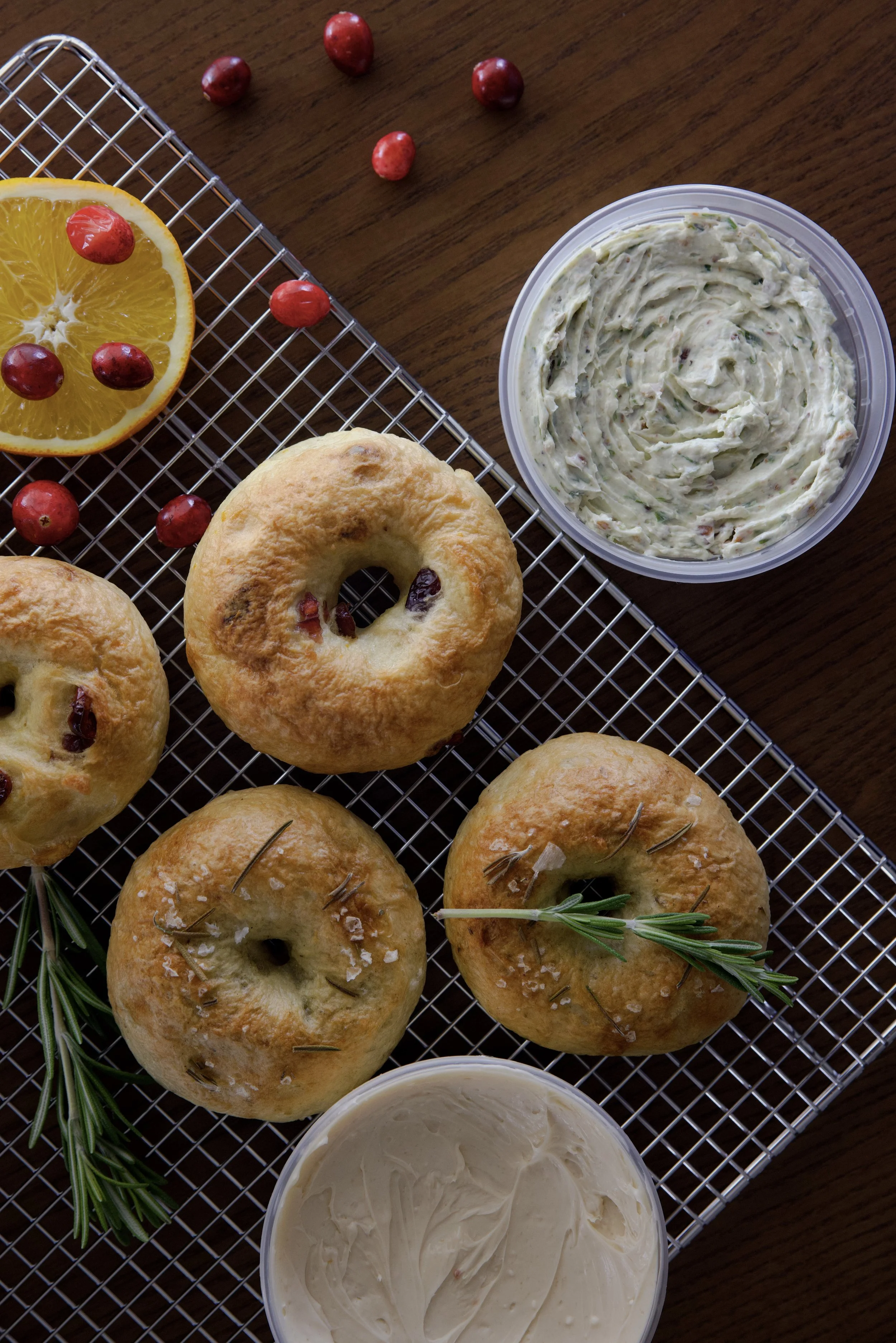 Four freshly baked bagels garnished with rosemary and coarse salt, served with a side of cream cheese, along with sliced orange and red berries, all arranged on a wire rack.