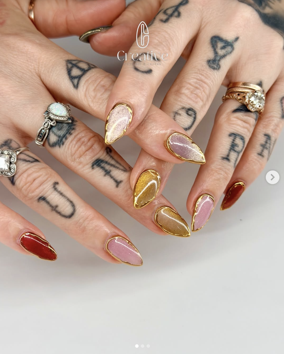 Hands with colorful, shiny, almond-shaped nails and various rings, including a large opal, gold accents, and a bright diamond, covered in tattoos of letters and symbols.