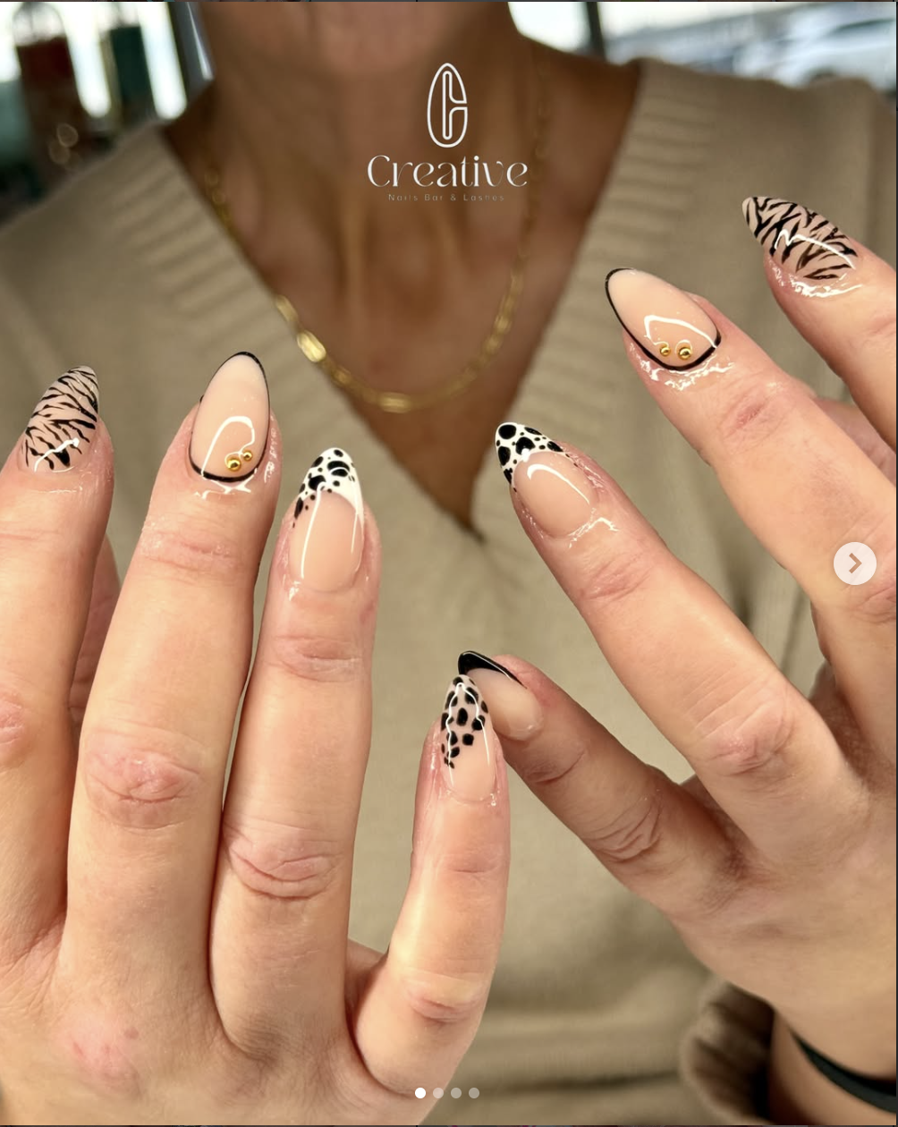 Close-up of a woman's hands with manicured nails featuring animal print designs in black, beige, and white, with gold and black accents, held up in front of a beige shirt and a blurred background.