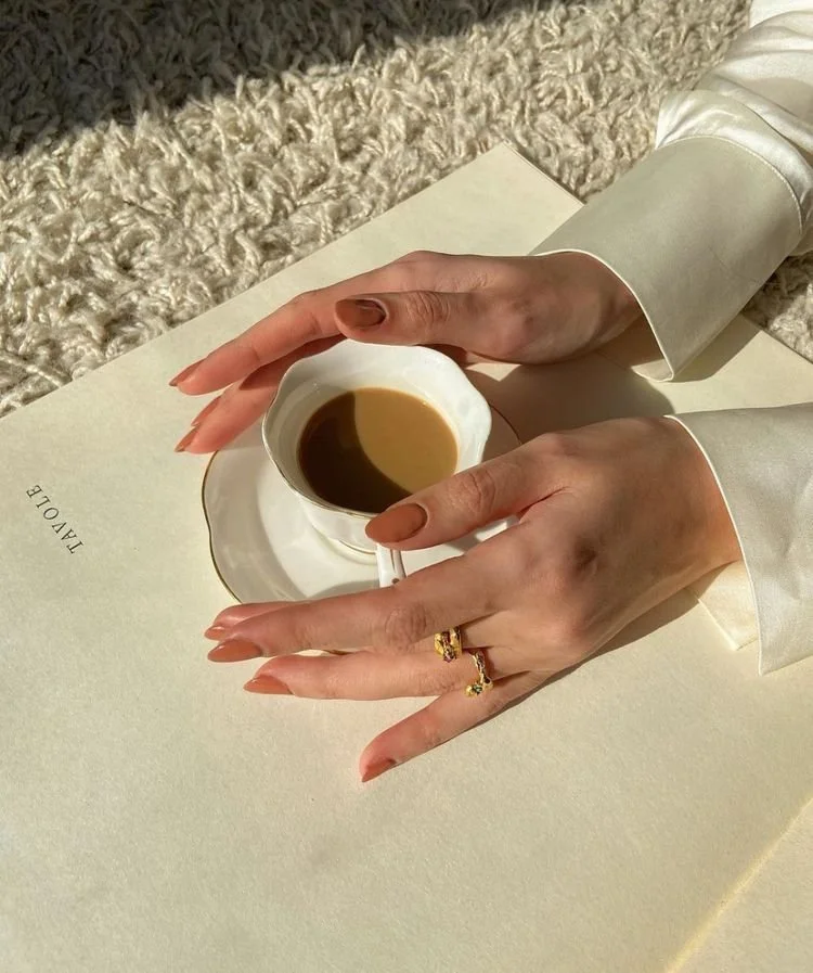 A person's hands holding a white teacup filled with coffee, with a ring on one finger. The hands rest on a cream-colored surface with the words 'IMAGO' printed on it. The background includes a plush, textured carpet and a silky, cream-colored sleeve.