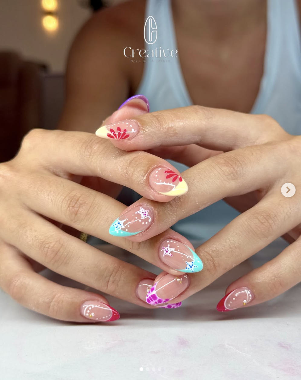 Colorful decorated nails with flower, star, and swirl designs in pink, yellow, blue, and purple on natural nails, held together by a person's hands with a blurred background and a logo overlay.