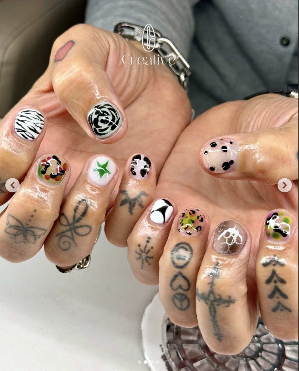 Close-up of hands with tattoos and colorful nail art featuring various patterns including zebra stripes, roses, polka dots, and abstract designs.