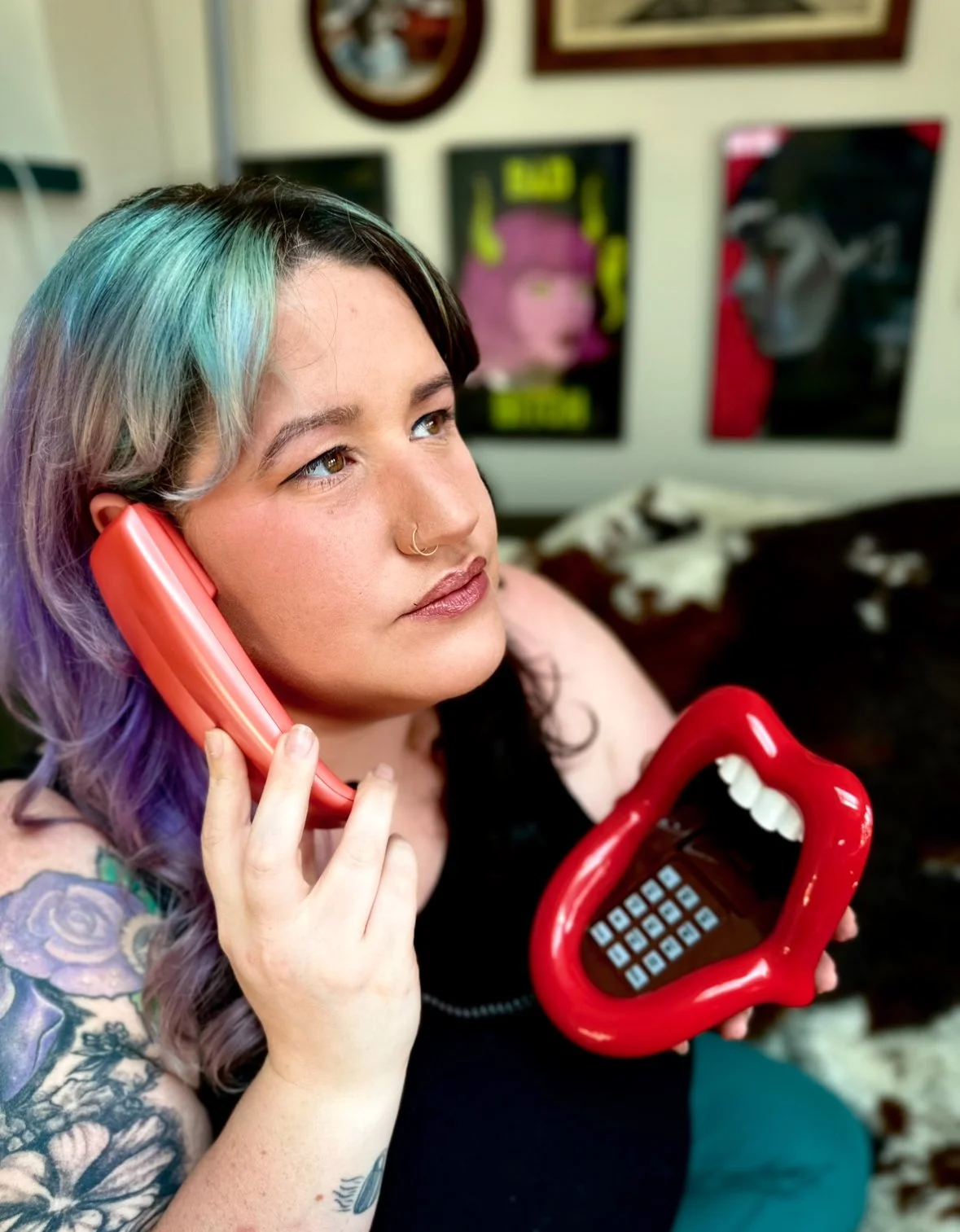 A woman with colorful hair talking on a pink phone while holding a red phone-shaped object with teeth and a tongue, in a room decorated with posters.
