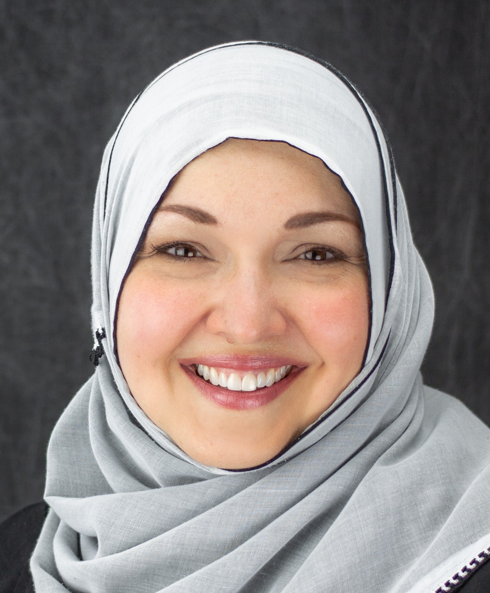 Smiling woman wearing a light gray hijab, black eyeliner, and lipstick, against a dark background. Dr. Sarah K. Samman serves zip codes 91344, 92131, and 92620.