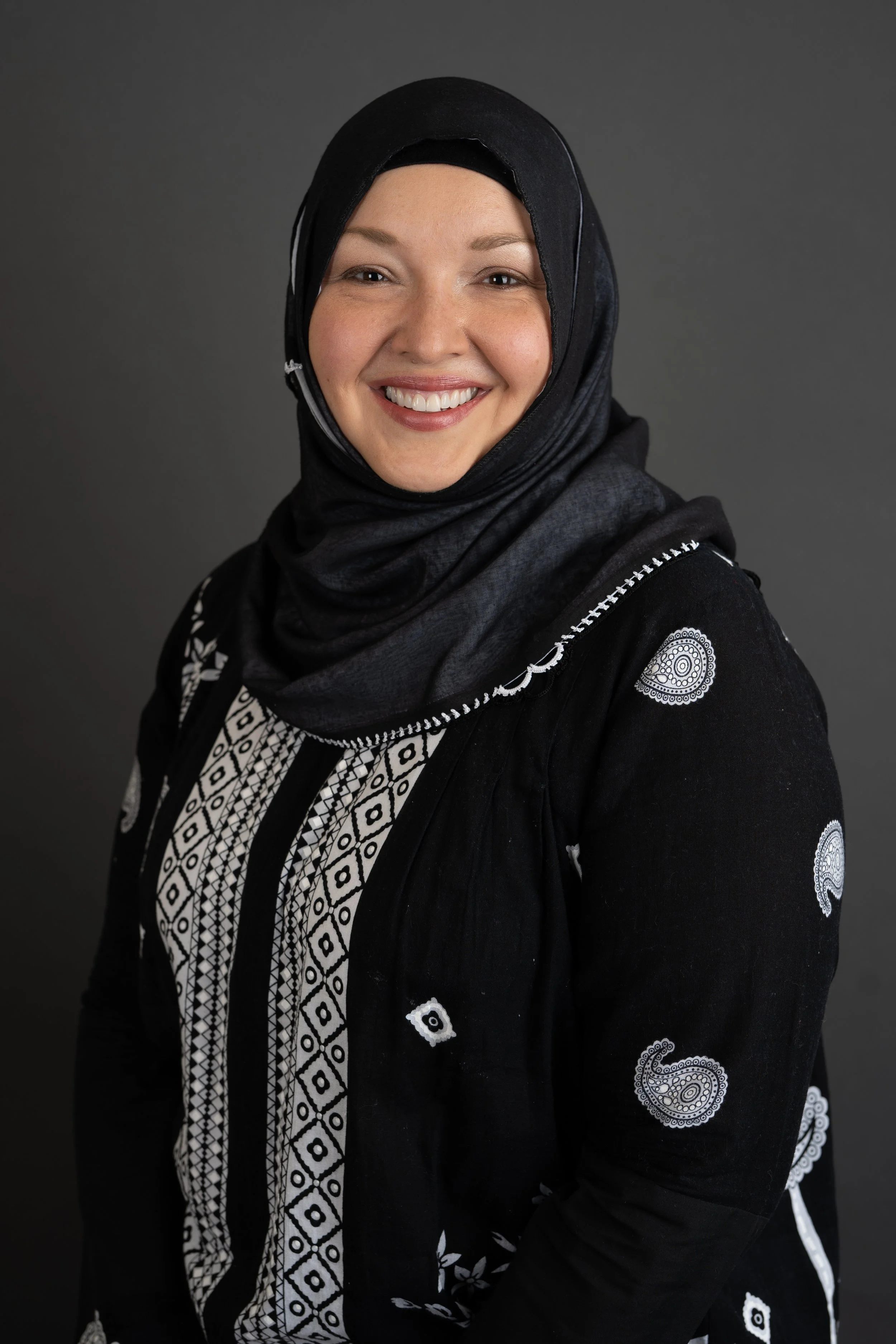 A smiling woman with light skin, wearing a black hijab and a black garment with white embroidered geometric and paisley patterns, standing against a dark gray background.