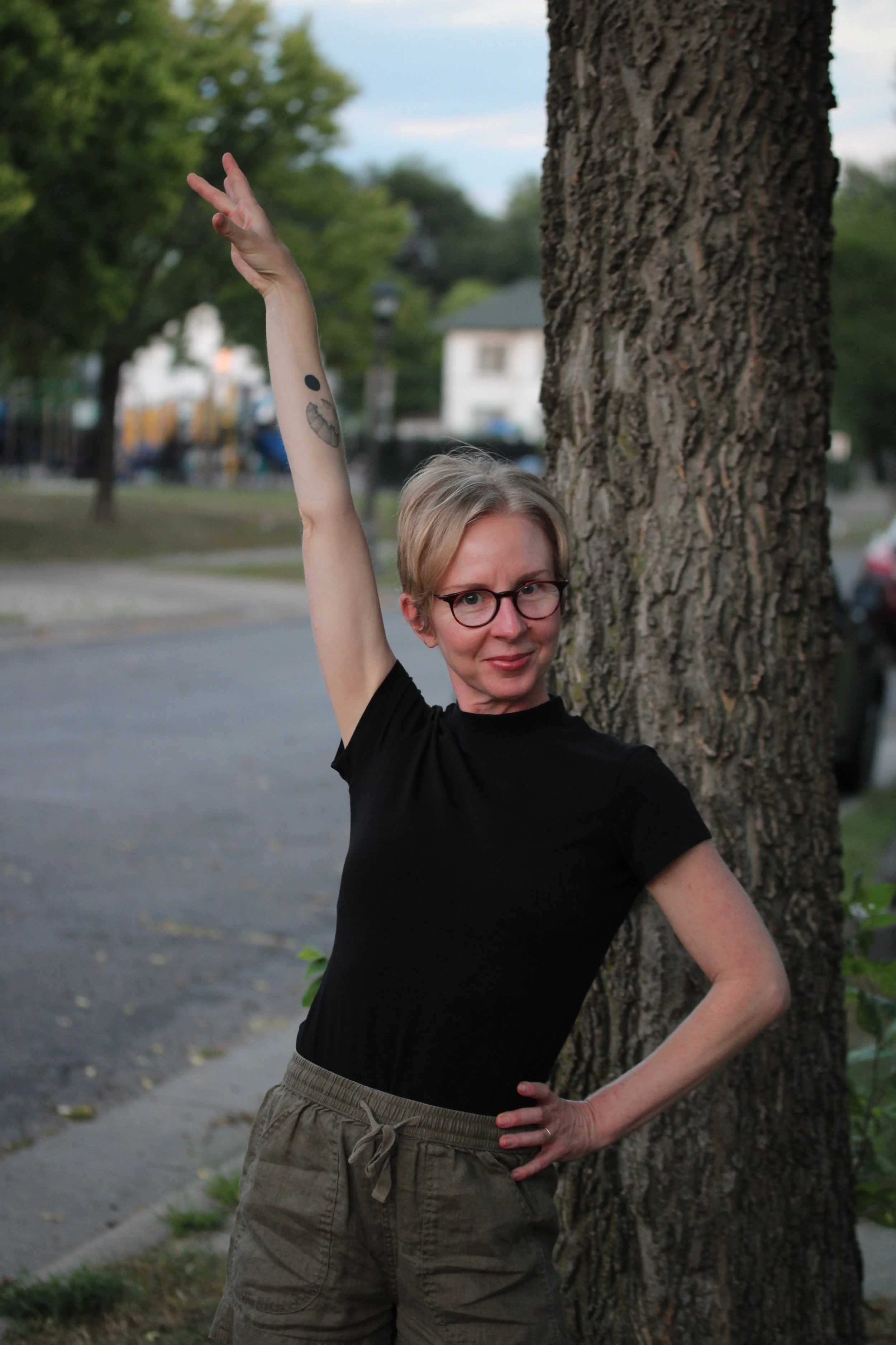 Rachel Kerr stands in front of a tree with one arm raised and the other on her hip. She is wearing glasses, a black shirt, olive green shorts, and has a circular tattoo on her raised arm.
