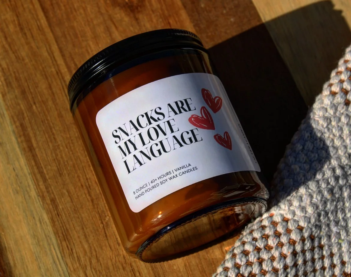 A candle jar labeled "Snacks are my love language" with red hearts on a wooden surface, partially resting on a textured cloth.