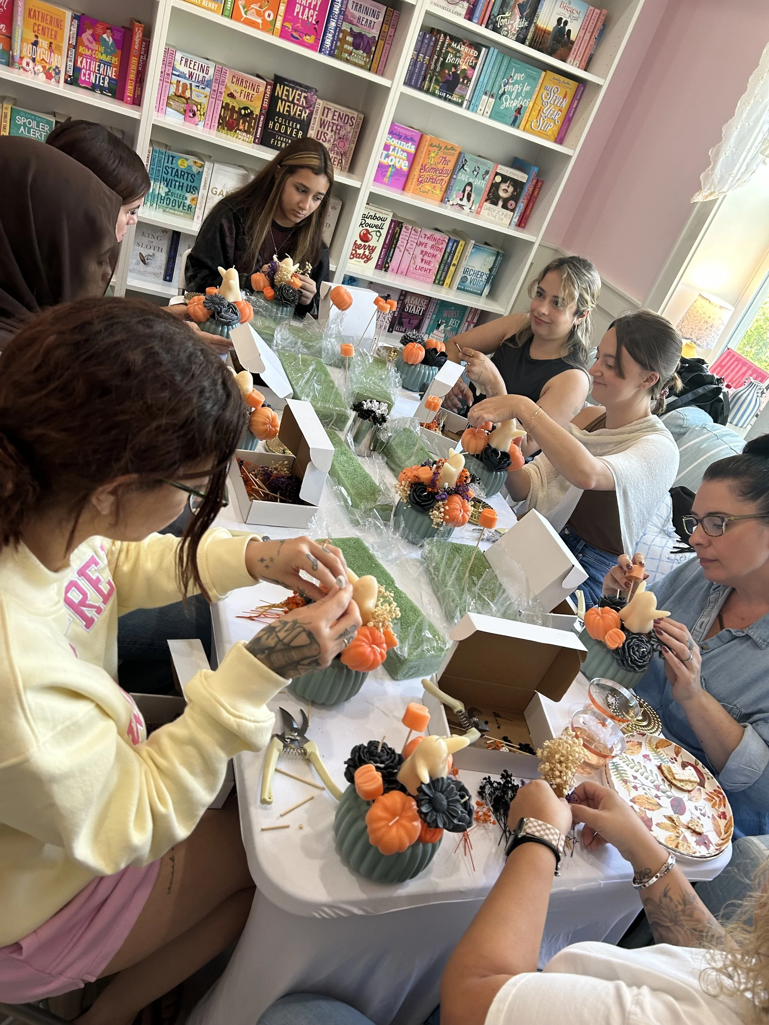 A group of women around a table creating Halloween-themed floral arrangements with pumpkins, black flowers, and gourds in teal containers, in a room with bookshelves filled with colorful books.