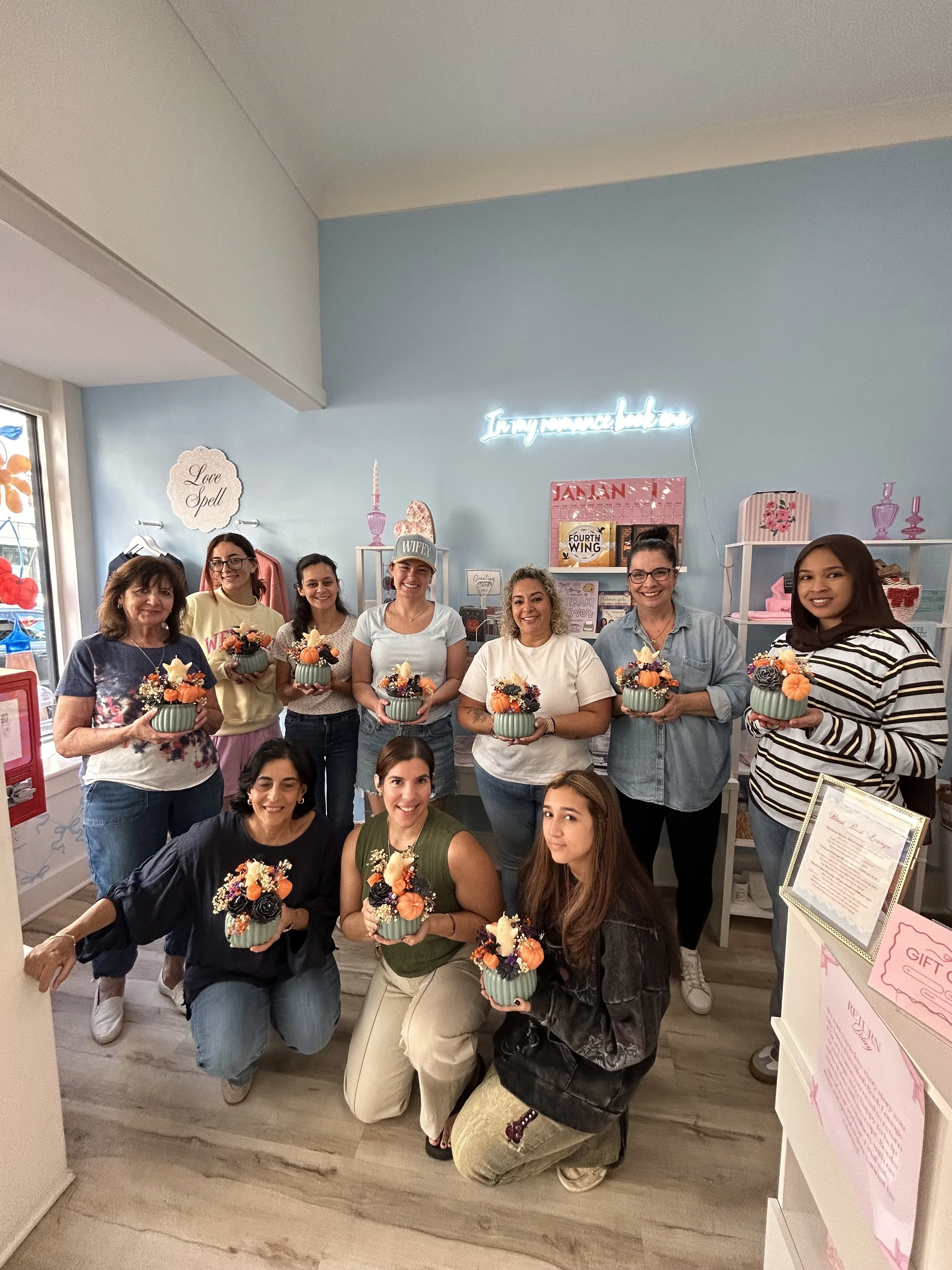 A group of women and girls holding small decorative flower arrangements in a boutique with light blue walls, shelves, and neon sign.