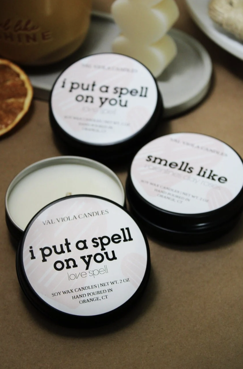 Three scented candles in small black tins labeled "I put a spell on you" by VAL VIOLA CANDLES, including one labeled "love spell" that is open and unlit, placed on a brown surface with decorative objects nearby.