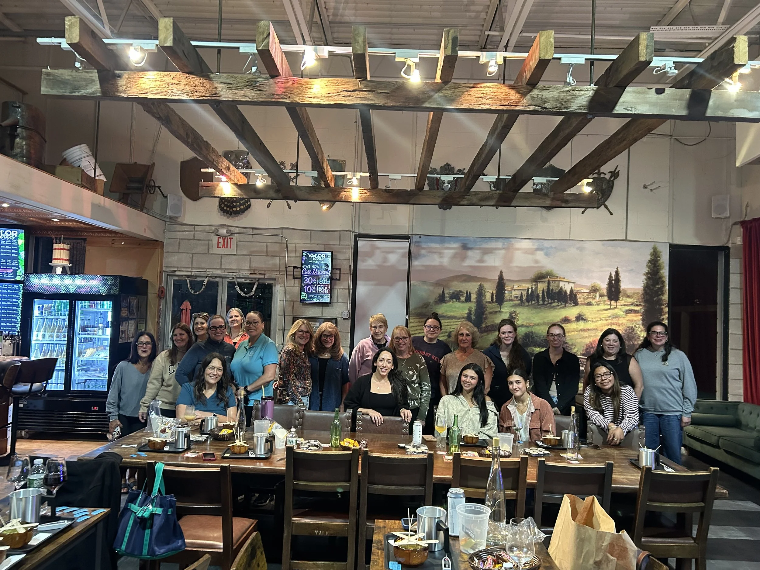 Group of people standing together around a dining table in a restaurant or banquet hall, with drinks and food remnants on the table, and a large landscape painting on the wall behind them.