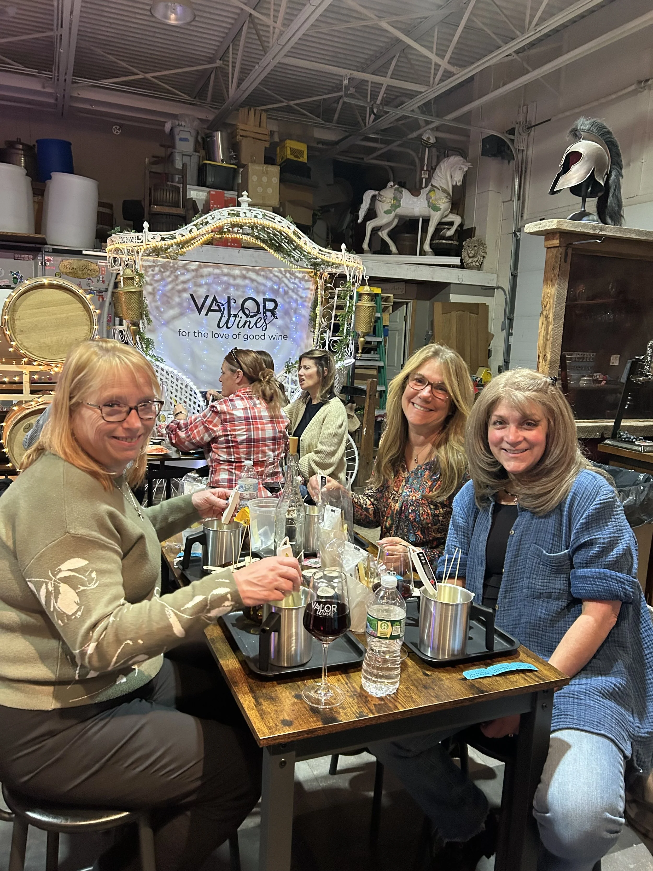Four women sitting around a candlelit table, enjoying wine and food in a cozy indoor setting with decorative elements and a sign that reads 'VALOR Wines' in the background.