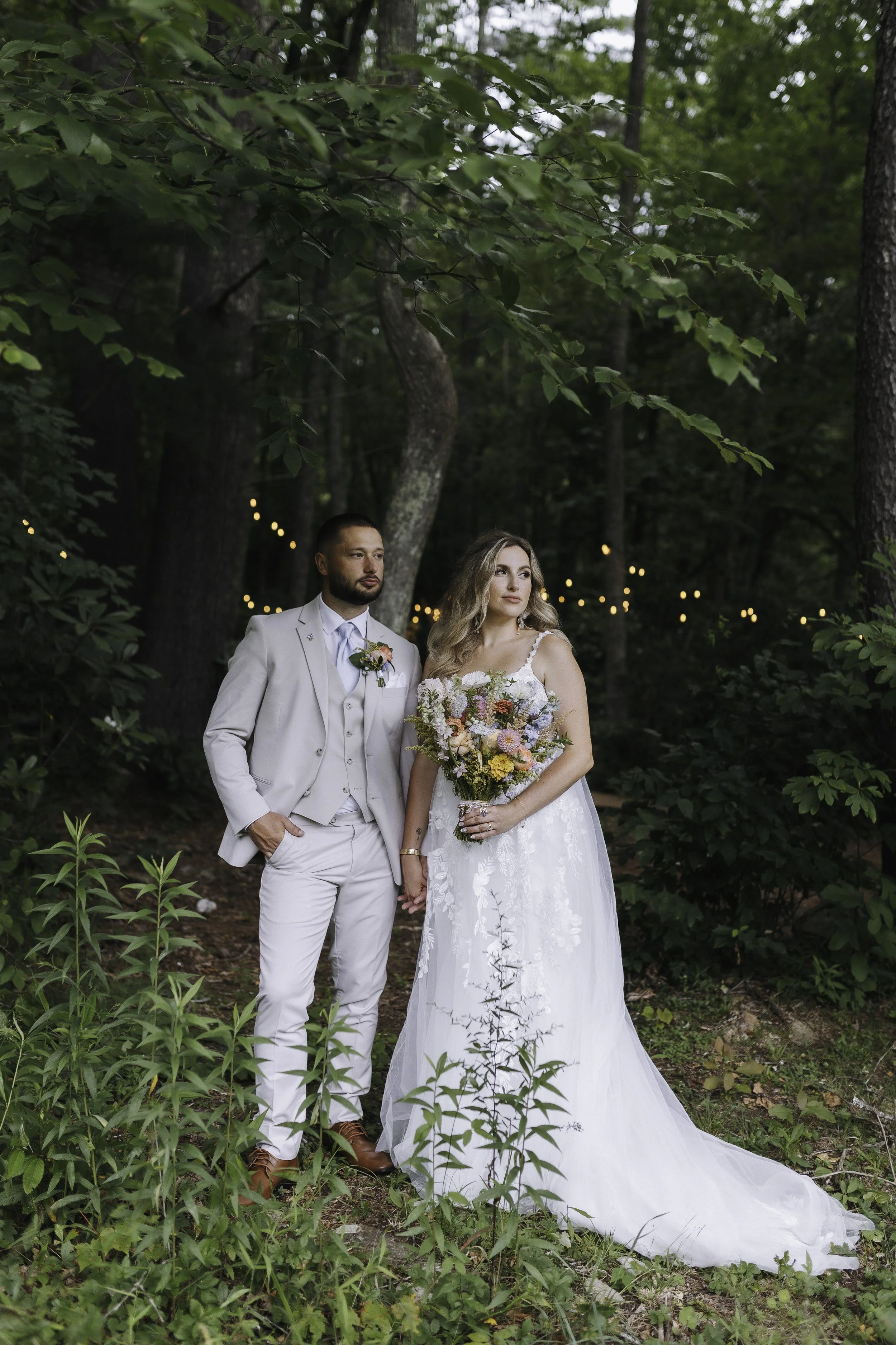 A bride and groom standing hand-in-hand in a lush green forest during a wedding photoshoot. The bride is holding a large bouquet of flowers, and both are dressed in formal wedding attire. The scene is decorated with string lights in the background.