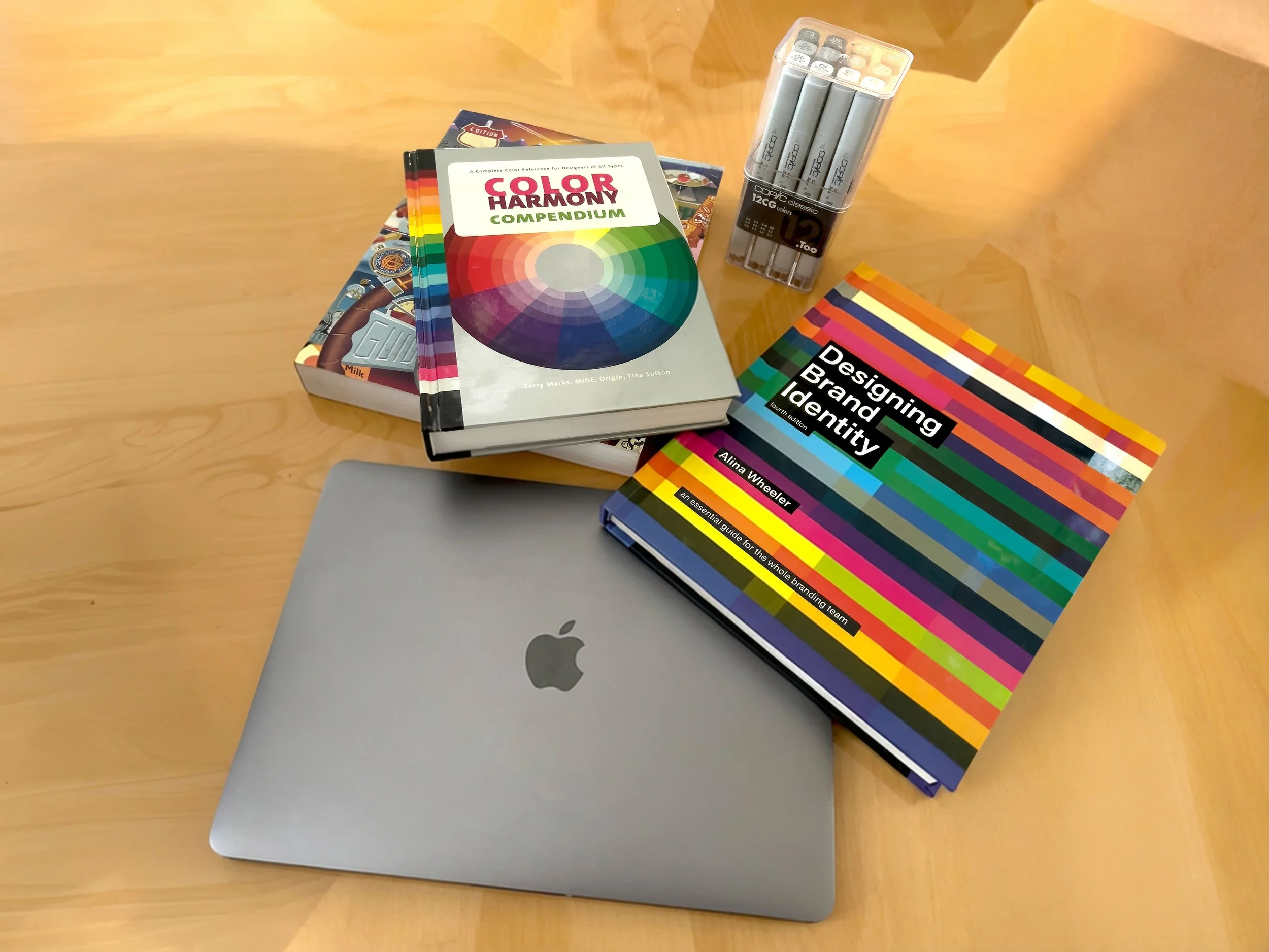 A MacBook laptop surrounded by color and branding books, highlighters, and a pack of 12 colored pens on a wooden table.