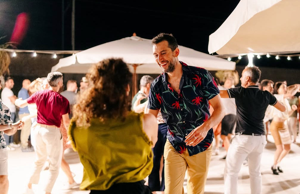 Persone che ballano e socializzano all'aperto di notte, sotto luci decorative, con un uomo sorridente al centro.