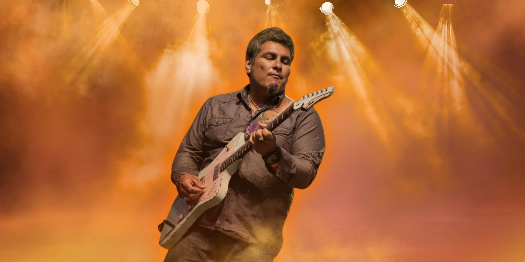 A man playing electric guitar on stage with orange lighting and spotlights. Jonn Del Toro Richardson.