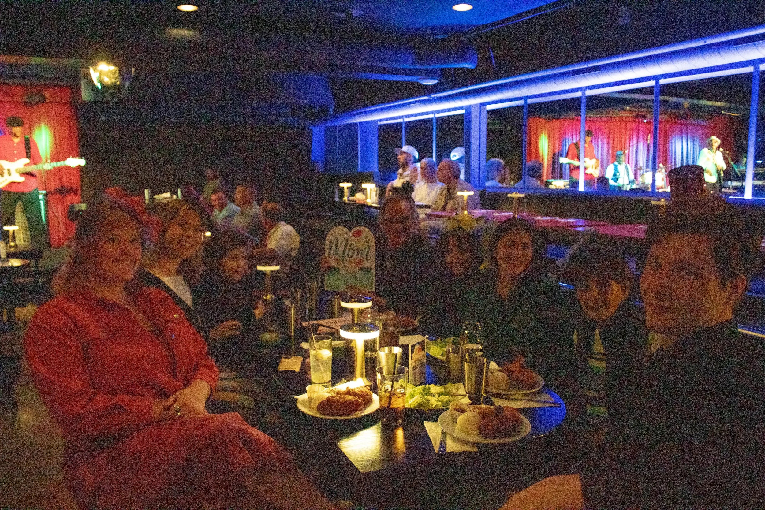 Group of people dining at a restaurant table with a stage and band in the background, low lighting, and neon accents.