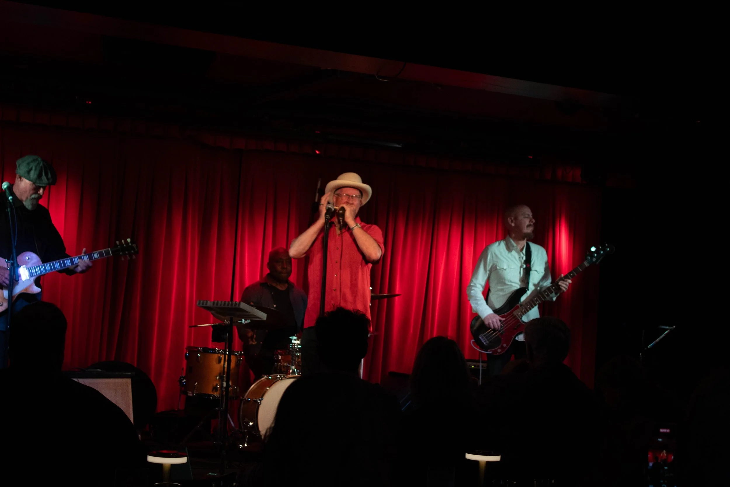A band performing on stage with four members; one playing guitar, another with a harmonica and wearing a hat, a drummer, and a bassist. The stage has red curtains and audience members are visible in the foreground.