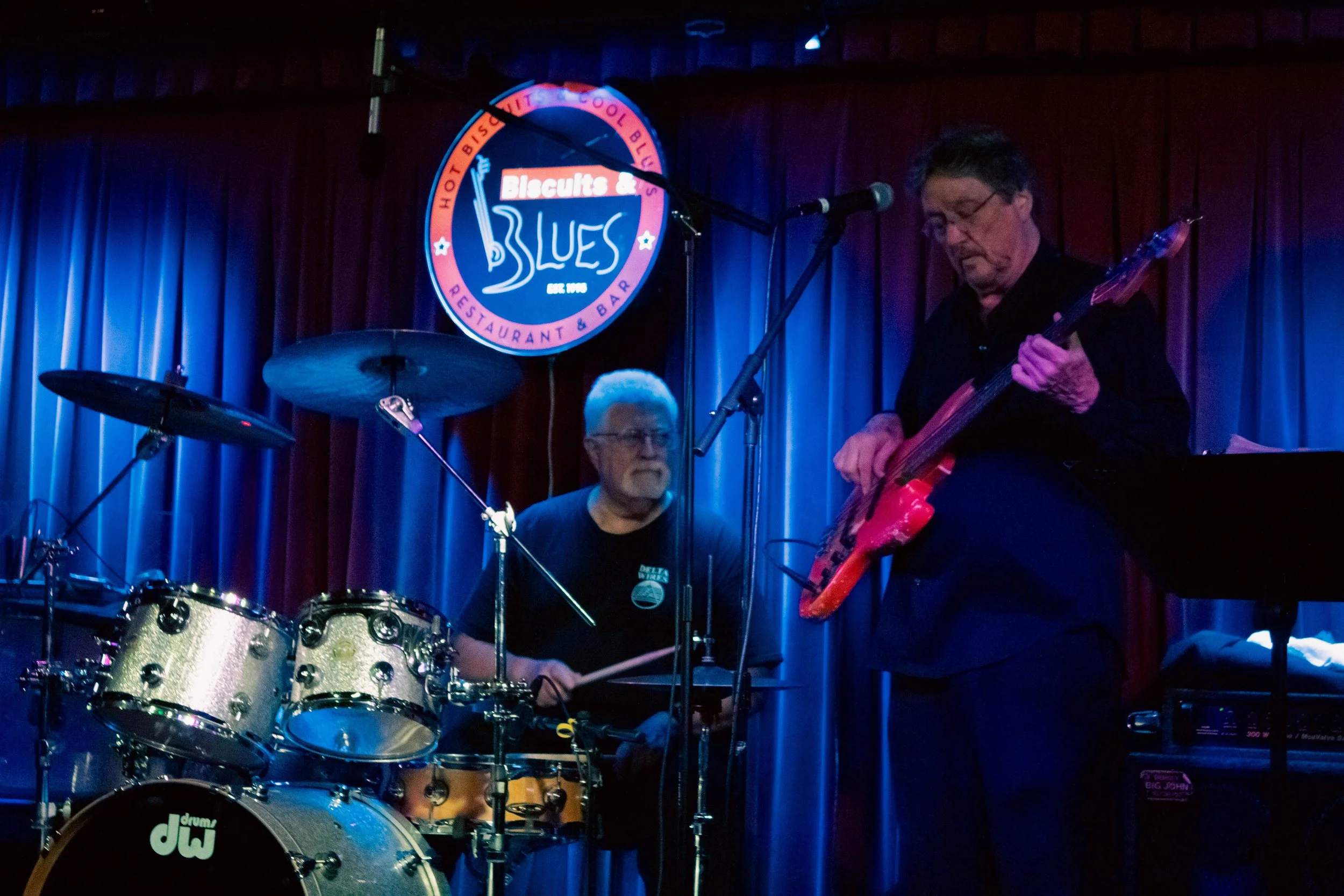 Two musicians performing on stage at a blues restaurant, one playing drums and the other playing bass guitar, with a blues-themed sign in the background.