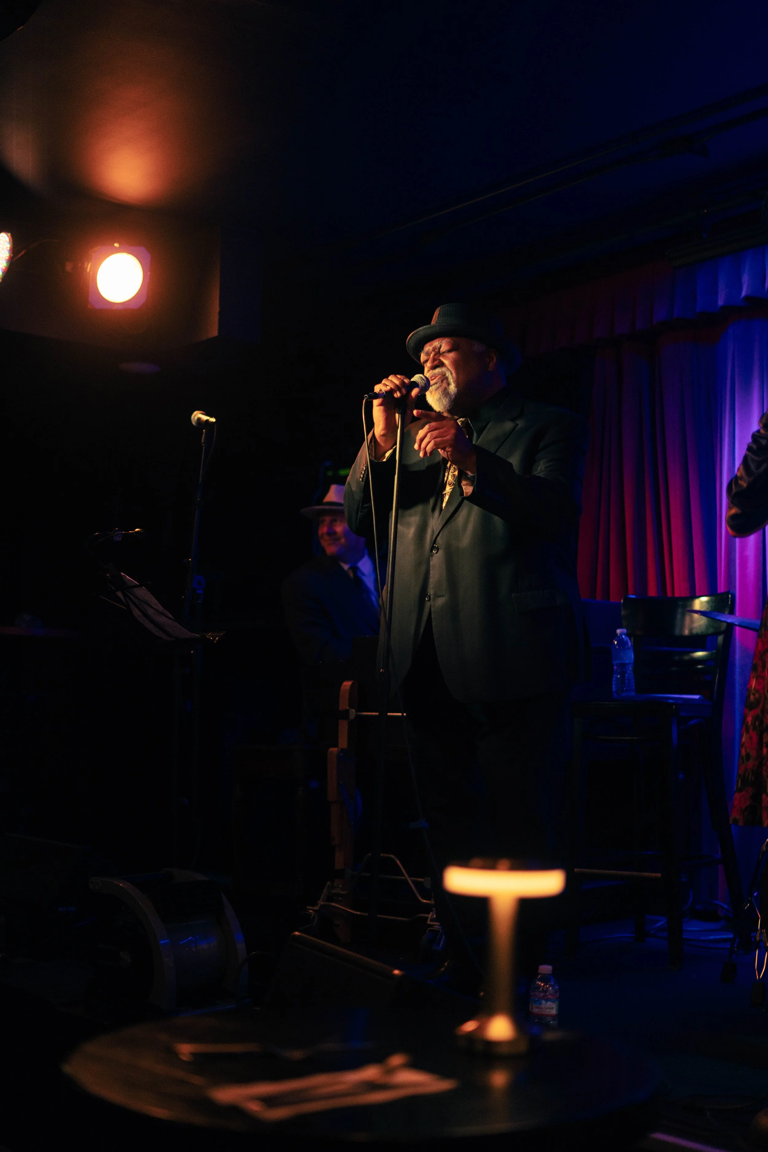 A man singing into a microphone on stage, wearing a black suit and hat, with purple curtains in the background, during a live performance in a dimly lit venue.
