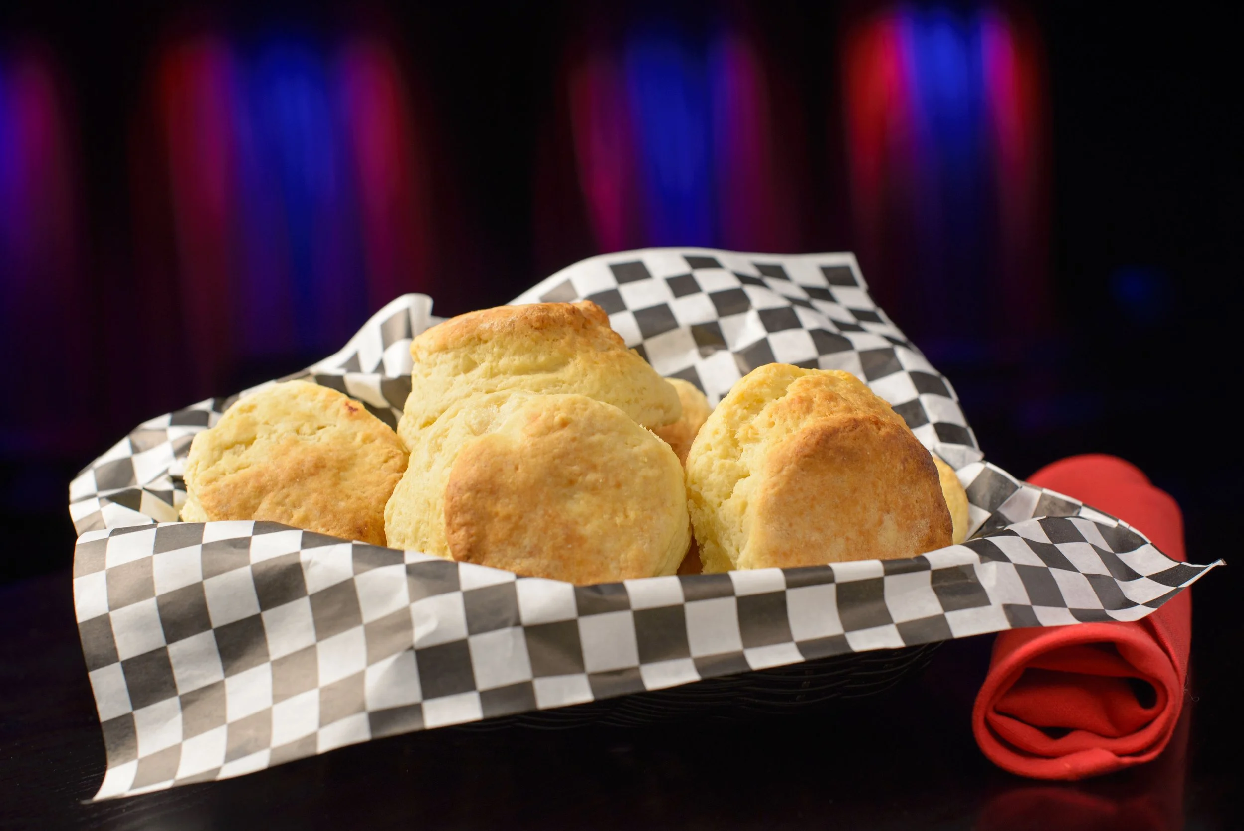Basket of golden-brown cornbread muffins on checkered paper with a red napkin underneath, against a dark background with red and blue lights.