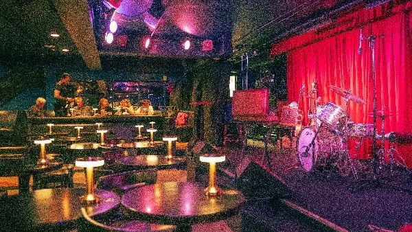 Empty stage with drums, piano, and microphone, red curtains, and an audience area with tables and stools in a music venue.