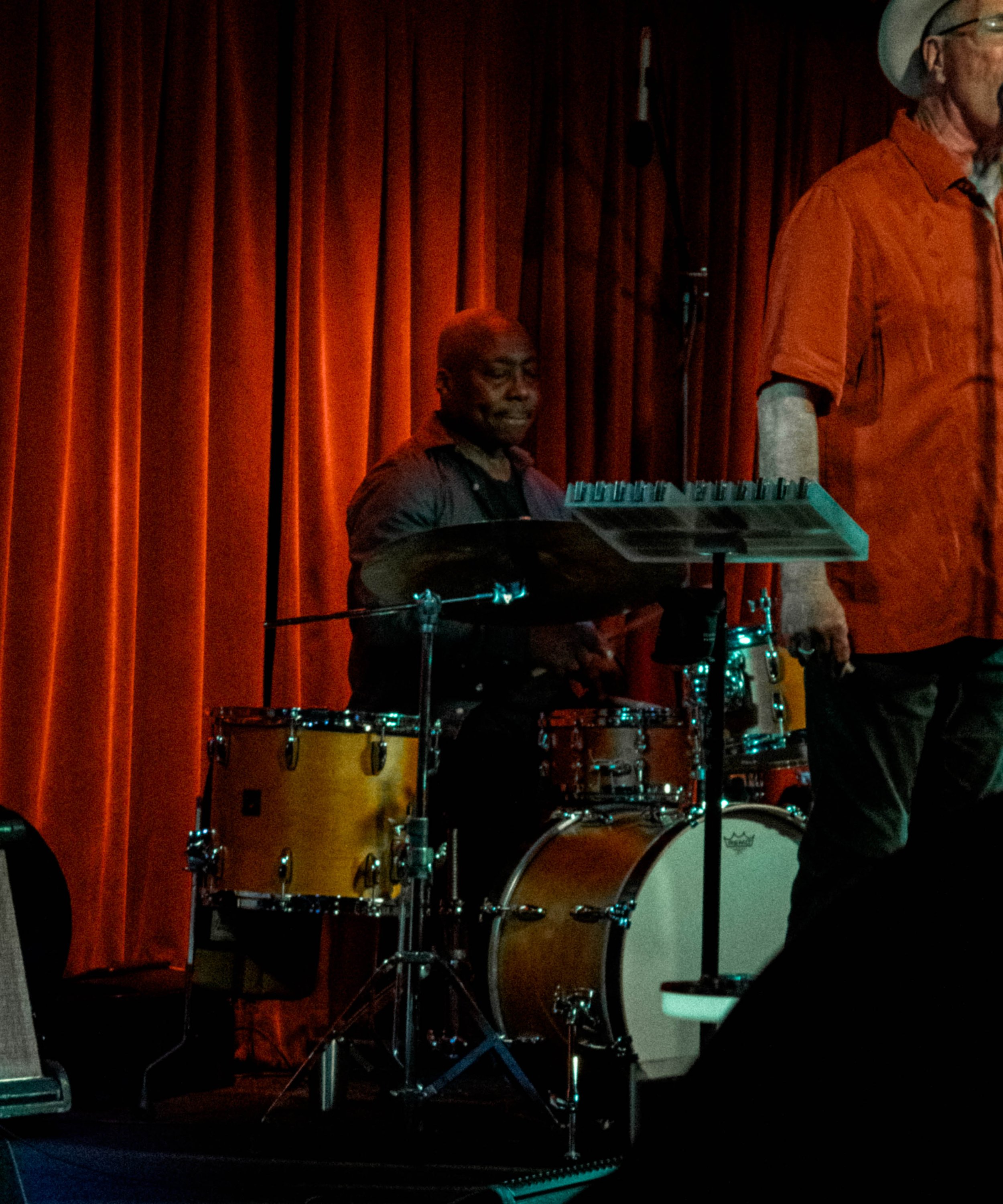 A person sitting behind a drum set on a stage with red curtains, playing the drums during a performance.