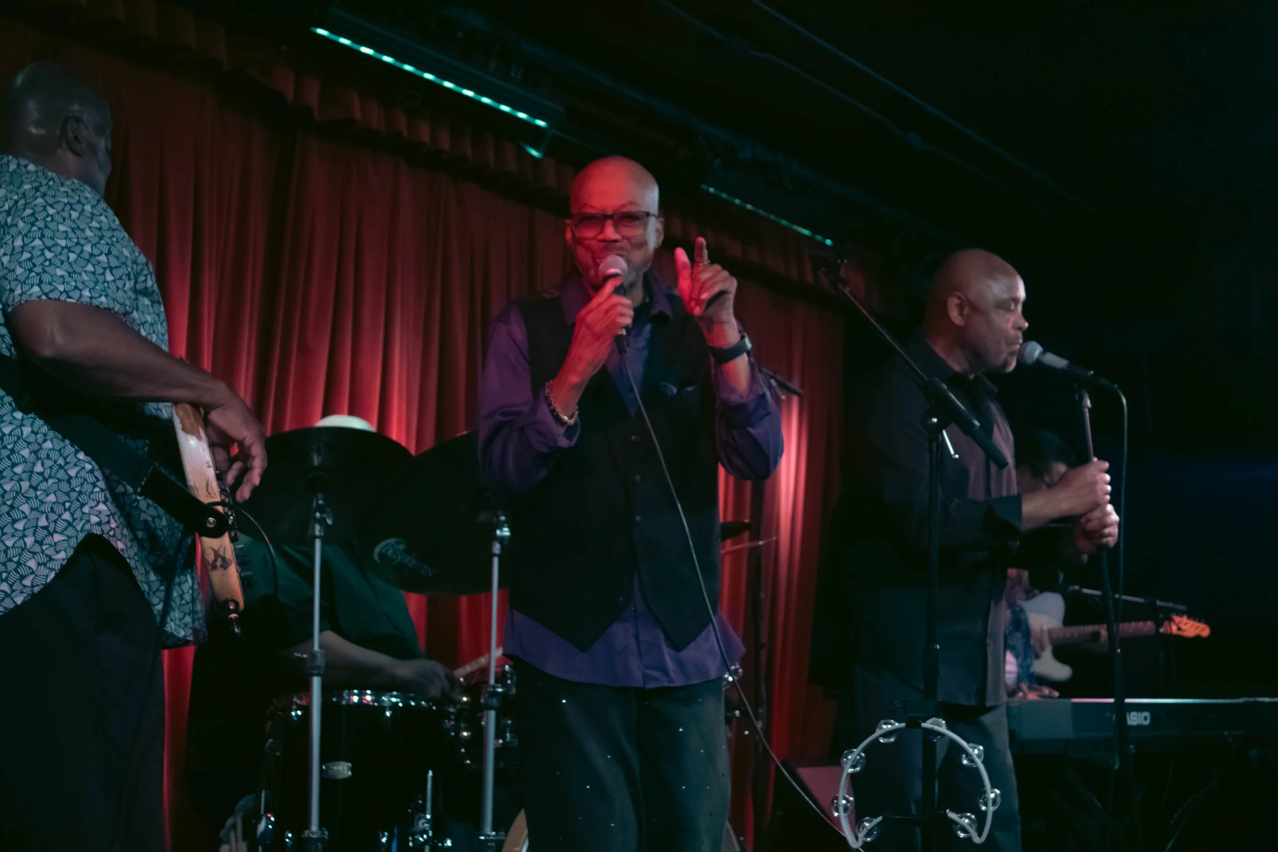 A live musical performance featuring a band with a singer, guitarist, drummer, and keyboardist on stage with a red curtain backdrop, dim lighting, and music equipment.