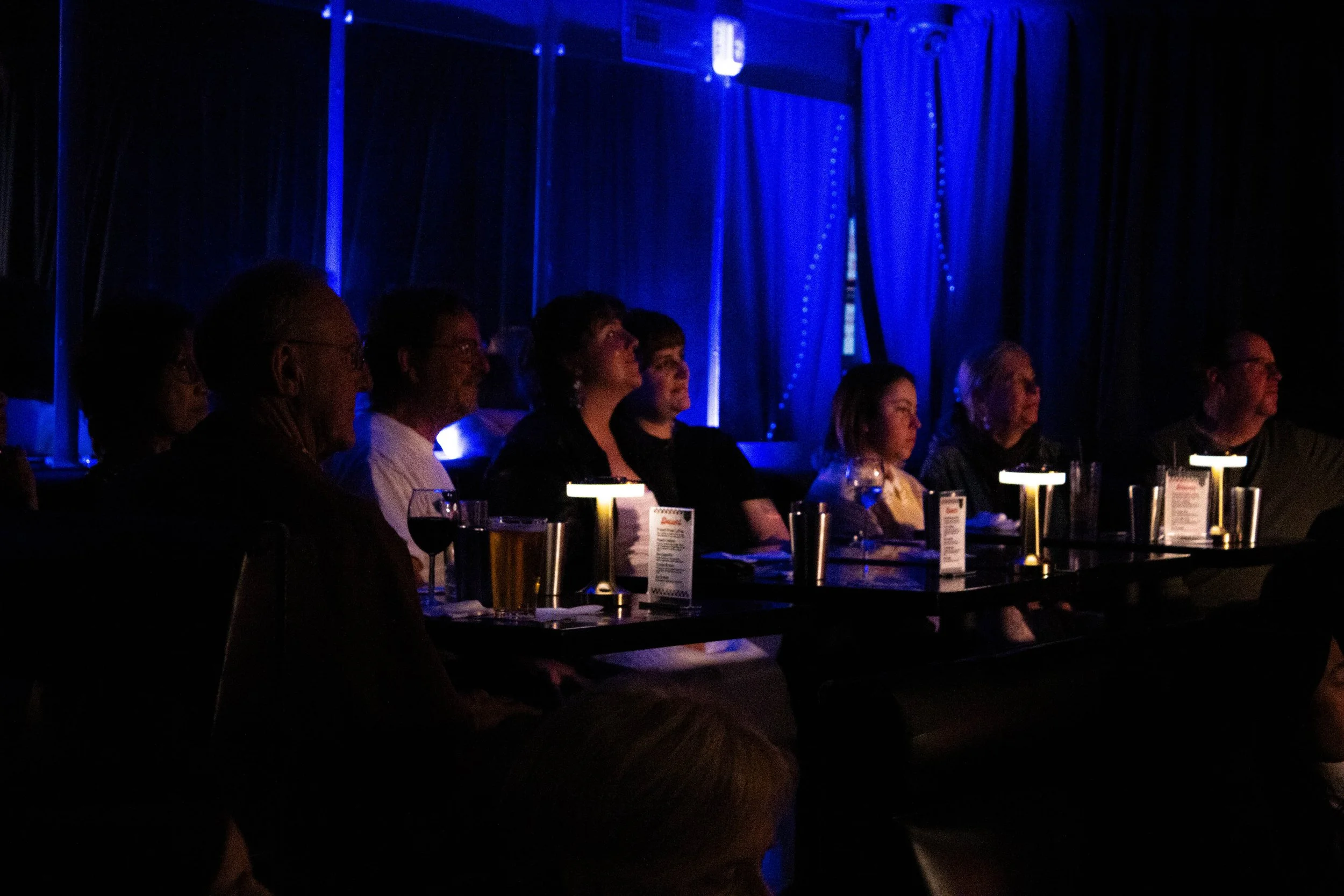 People sitting at a table in a dark theater or concert venue, attentively watching a performance, illuminated by dim blue lighting.