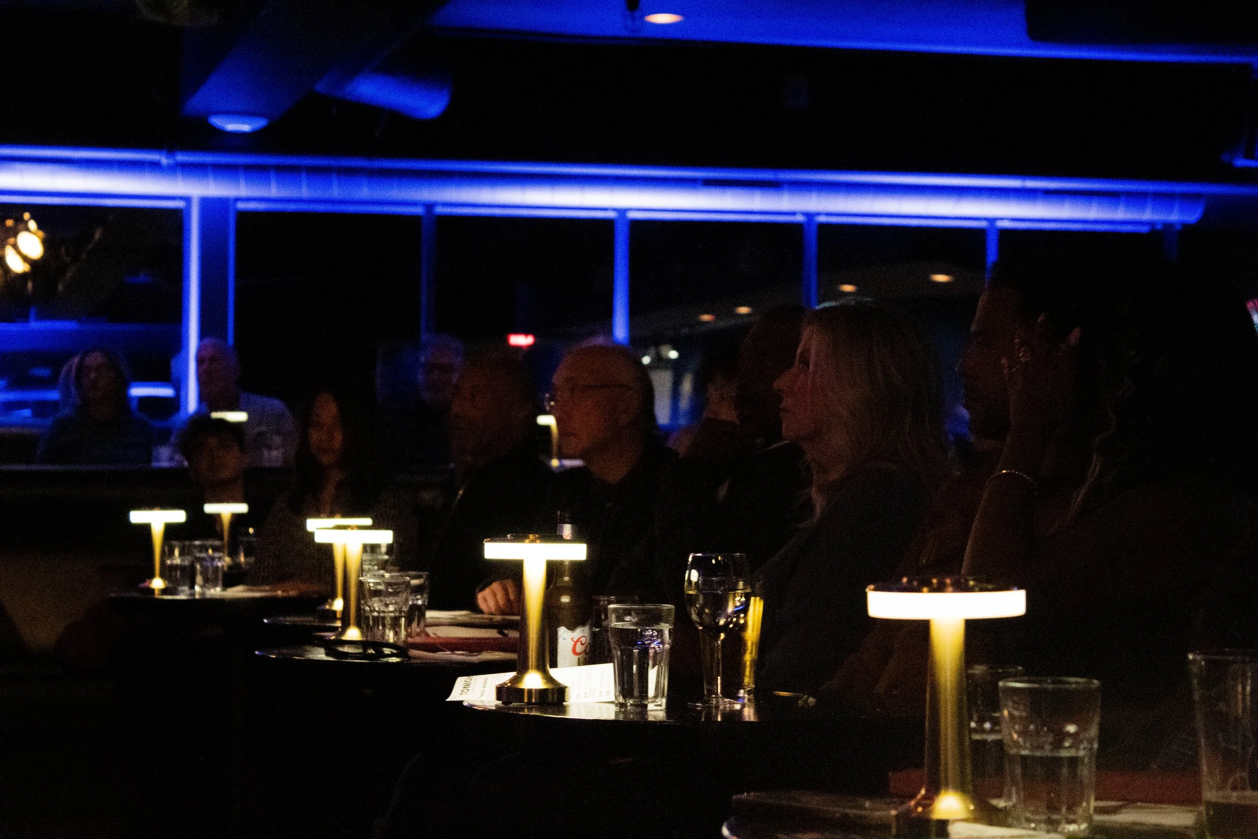 People sitting at a table with glasses of water and wine, illuminated by modern table lamps, in a dark indoor setting, possibly a restaurant or bar.